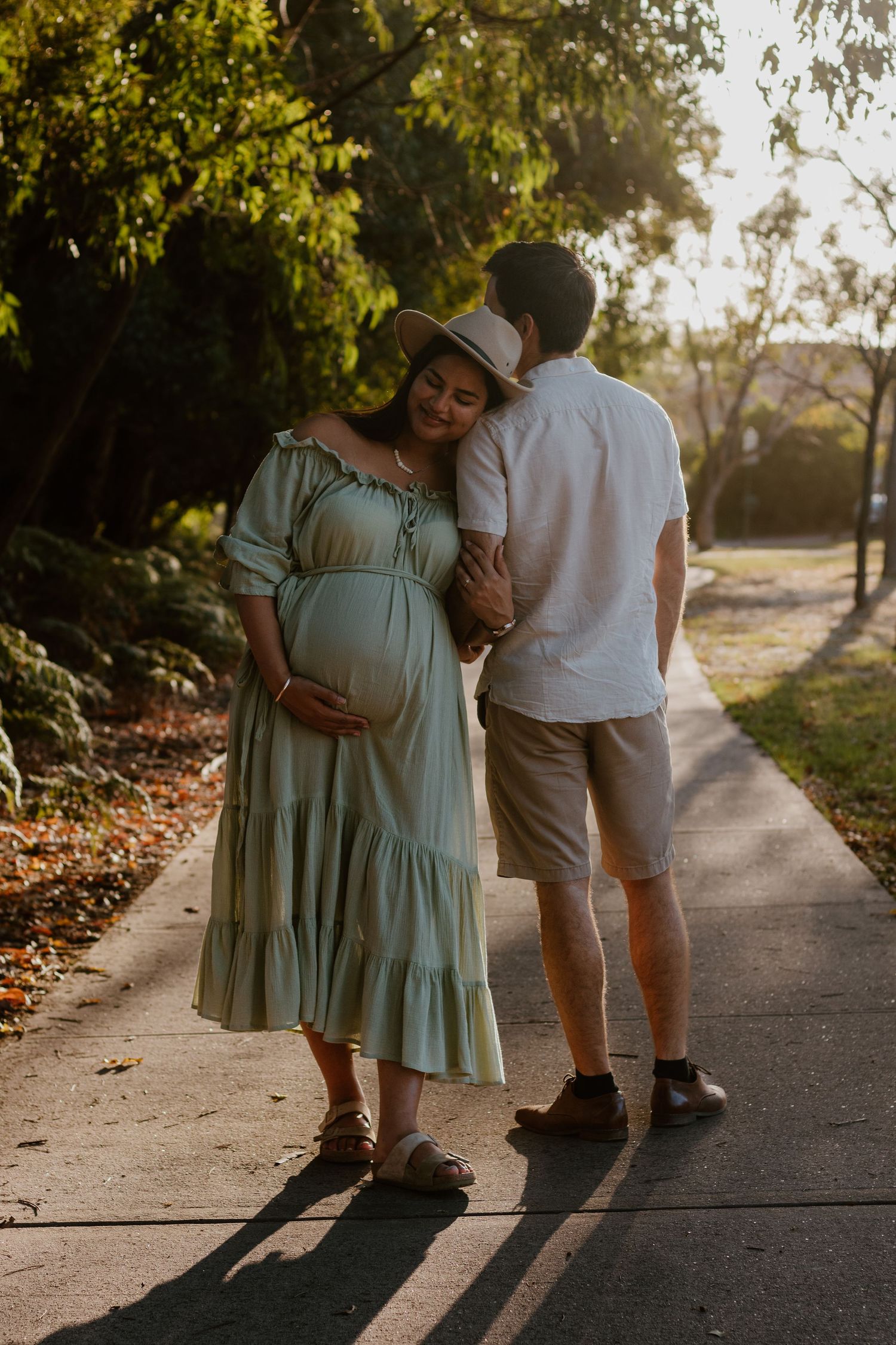Pregnant wife holding her belly with one hand and her husband's arm with the other. They are standing on a beautiful street at golden hour