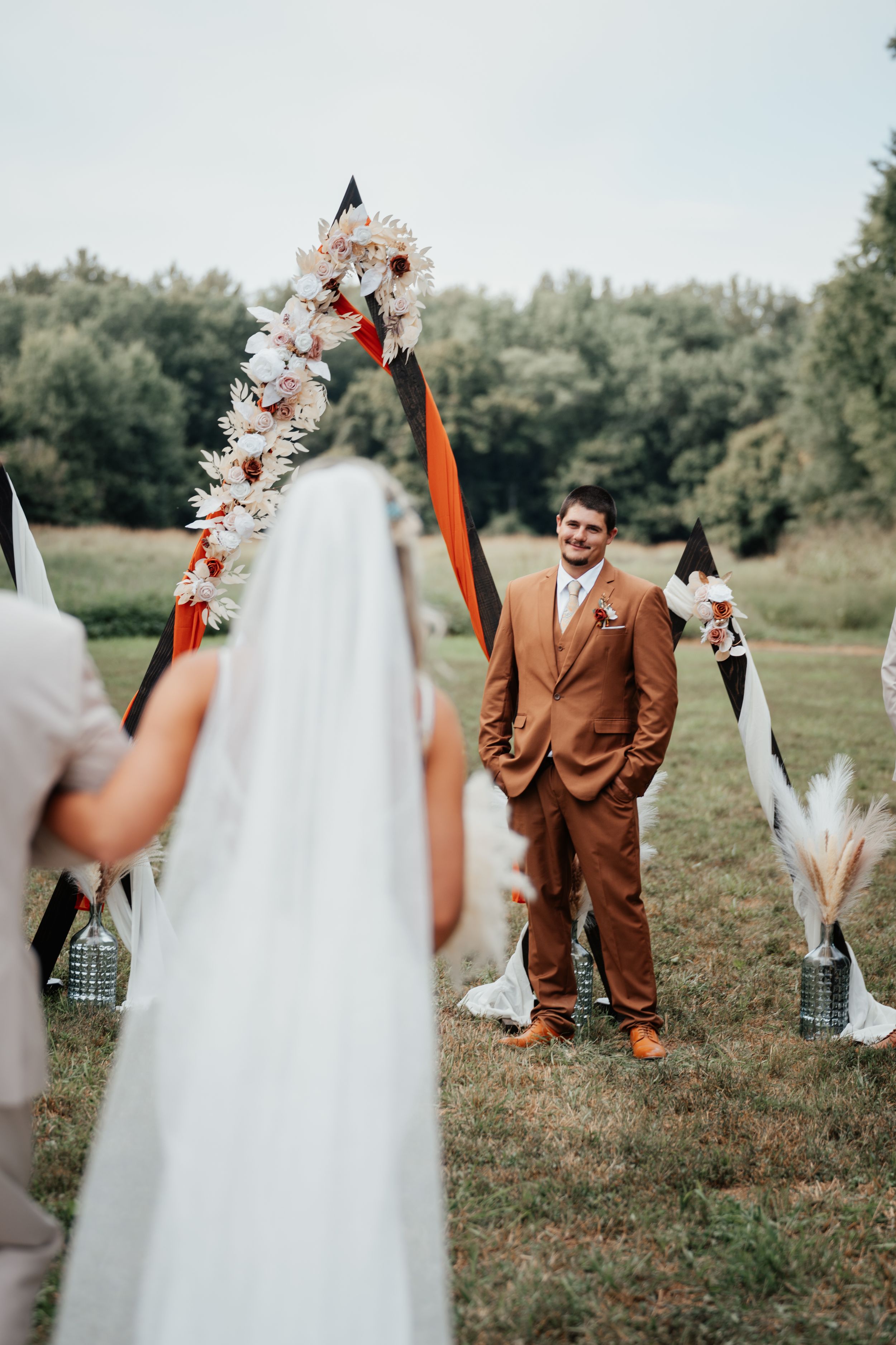 Couple at rustic outdoor wedding ceremony with floral arch and white draping in a grassy field.