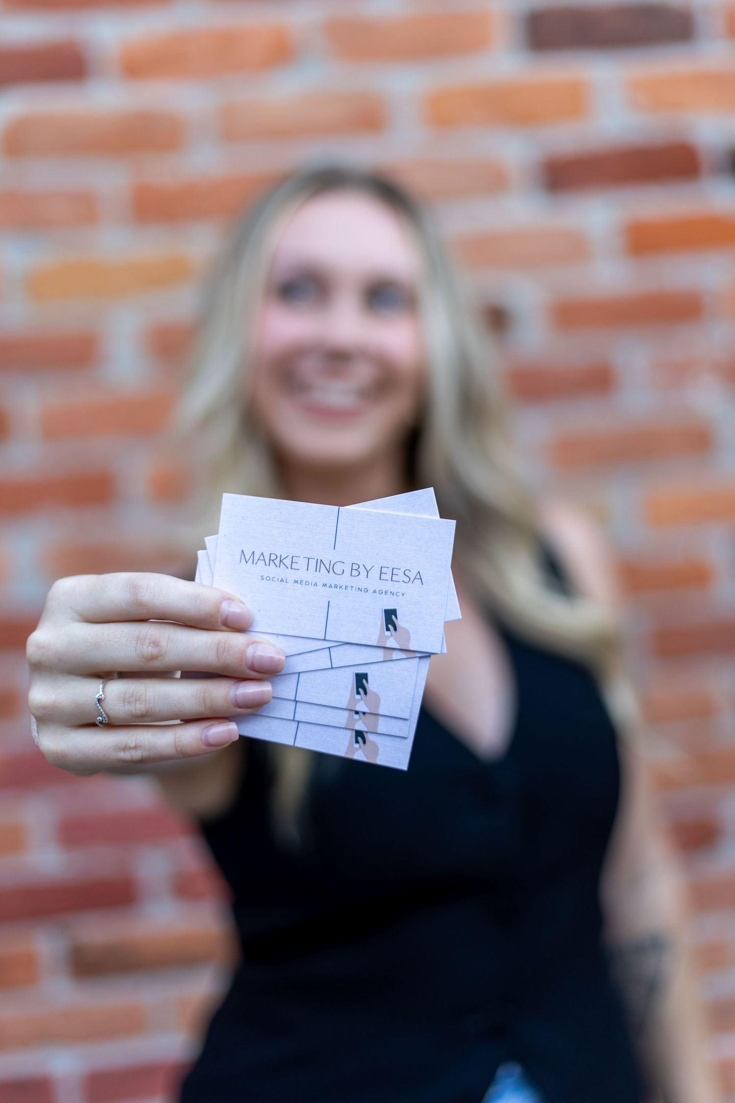 Smiling blonde woman holding Marketing by Eesa business cards in front of brick wall background.