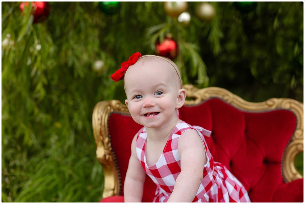 Smiling baby girl in red plaid dress with red bow sitting on velvet chair with Christmas tree backdrop.