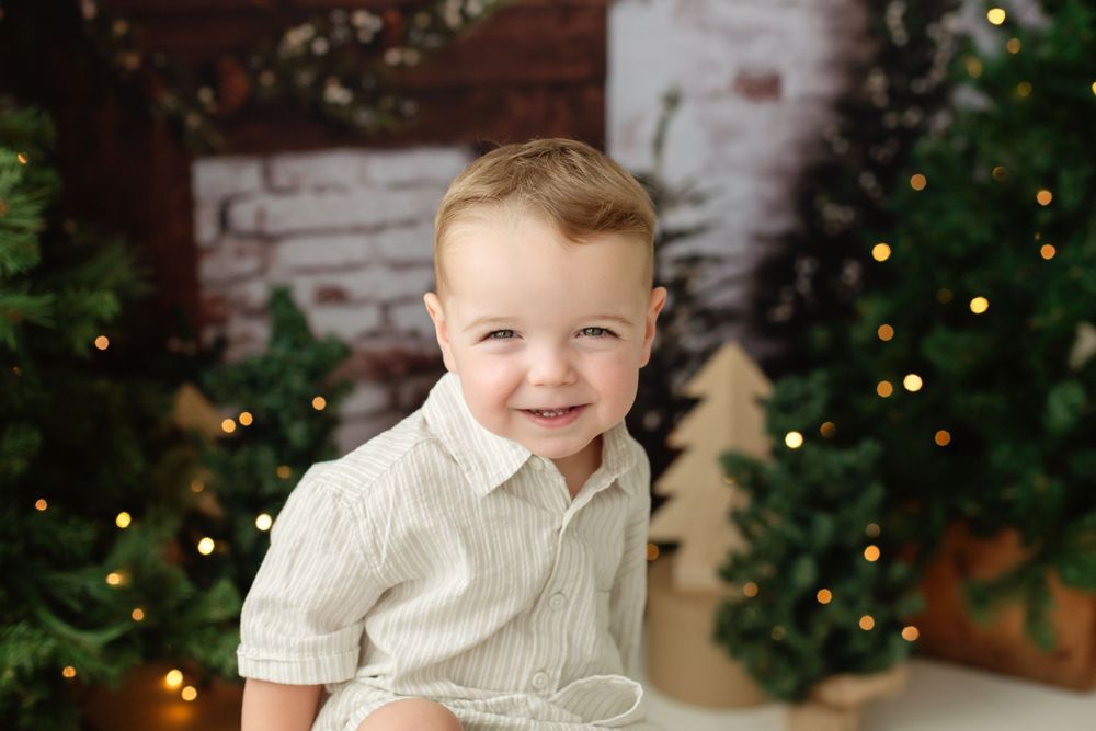 Adorable toddler boy in white shirt smiling in front of decorated Christmas trees with warm lights.