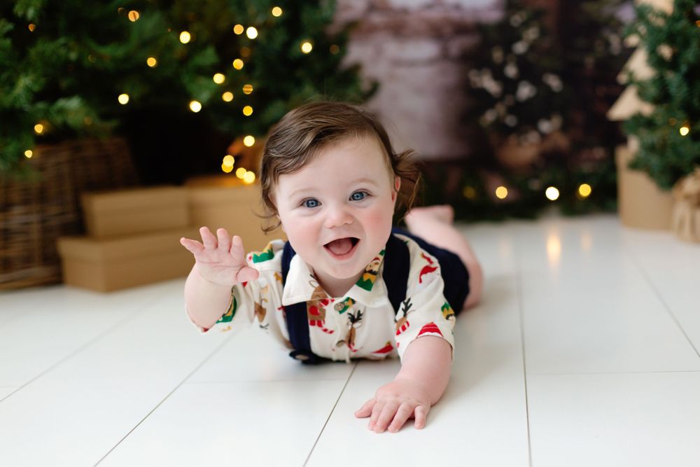 Smiling baby in festive romper lying on white floor with Christmas trees and lights in background.