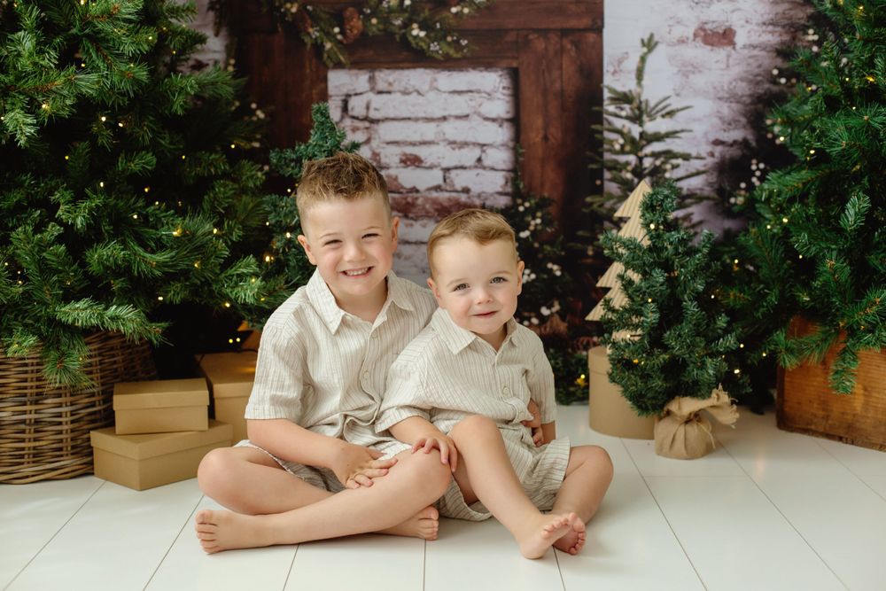 Two young brothers in matching outfits smiling together surrounded by Christmas trees and gifts.