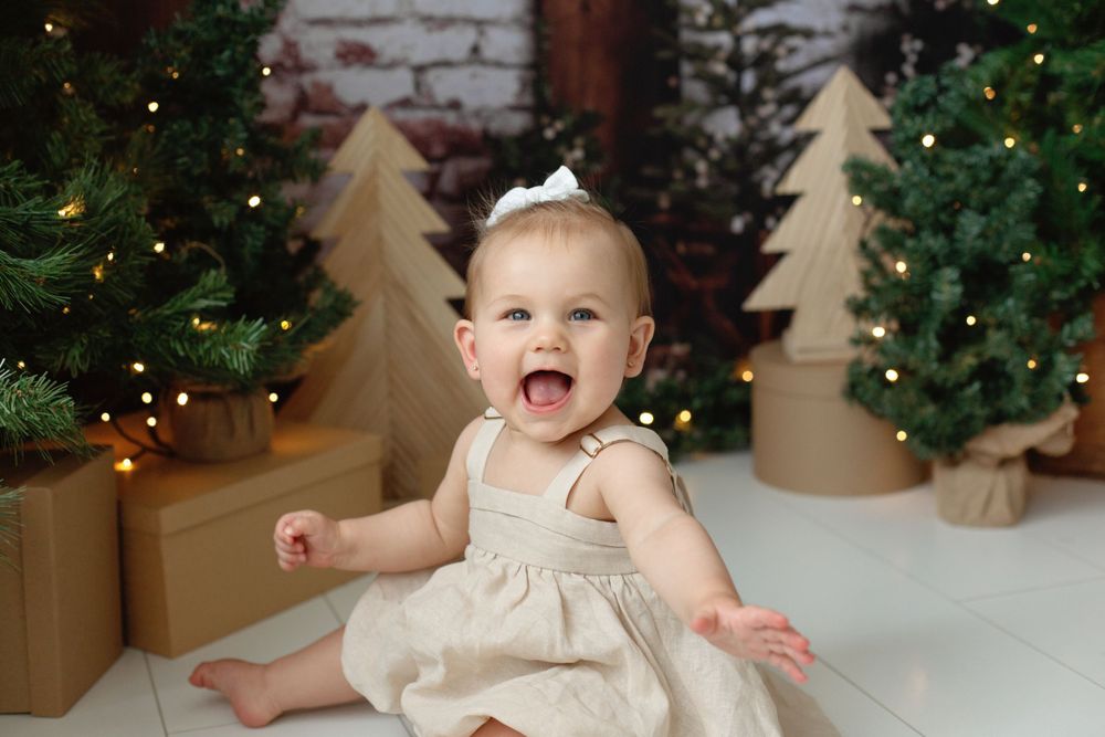 Happy baby girl in cream dress sitting among Christmas trees and gift boxes with bokeh lights.