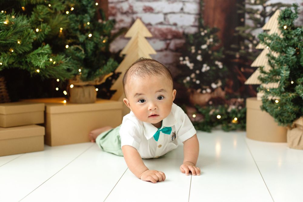 Asian baby boy in bow tie outfit crawling on white floor with Christmas trees and wooden decor.