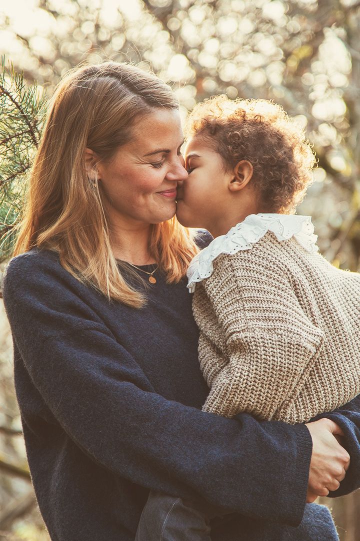 Dotter som ger sin mamma en puss på kinden i skogen, varmt fint ljus, jordnära färger, kärleksfull bild.