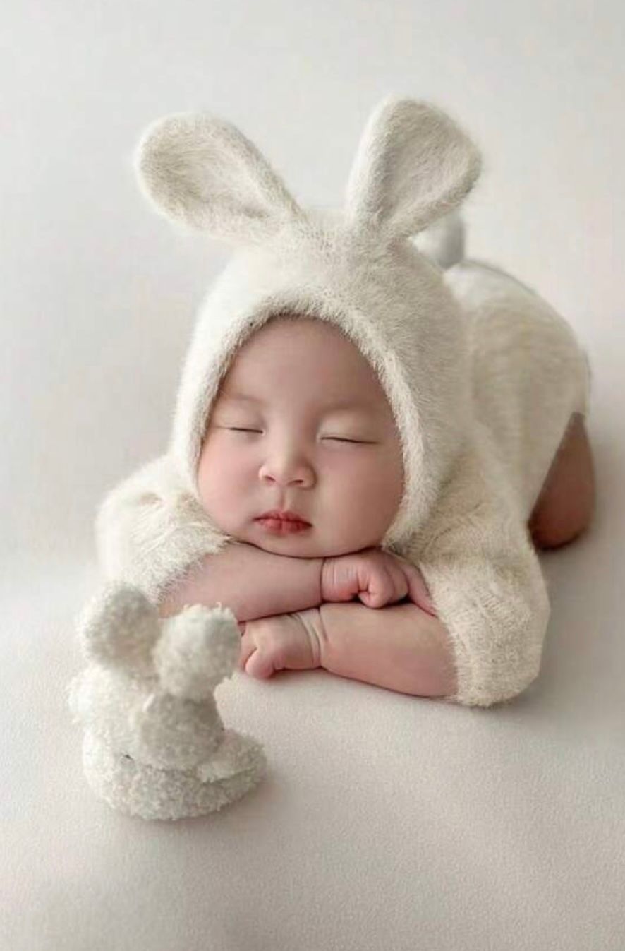 Sleeping newborn wrapped in soft white bunny-themed outfit on light background.