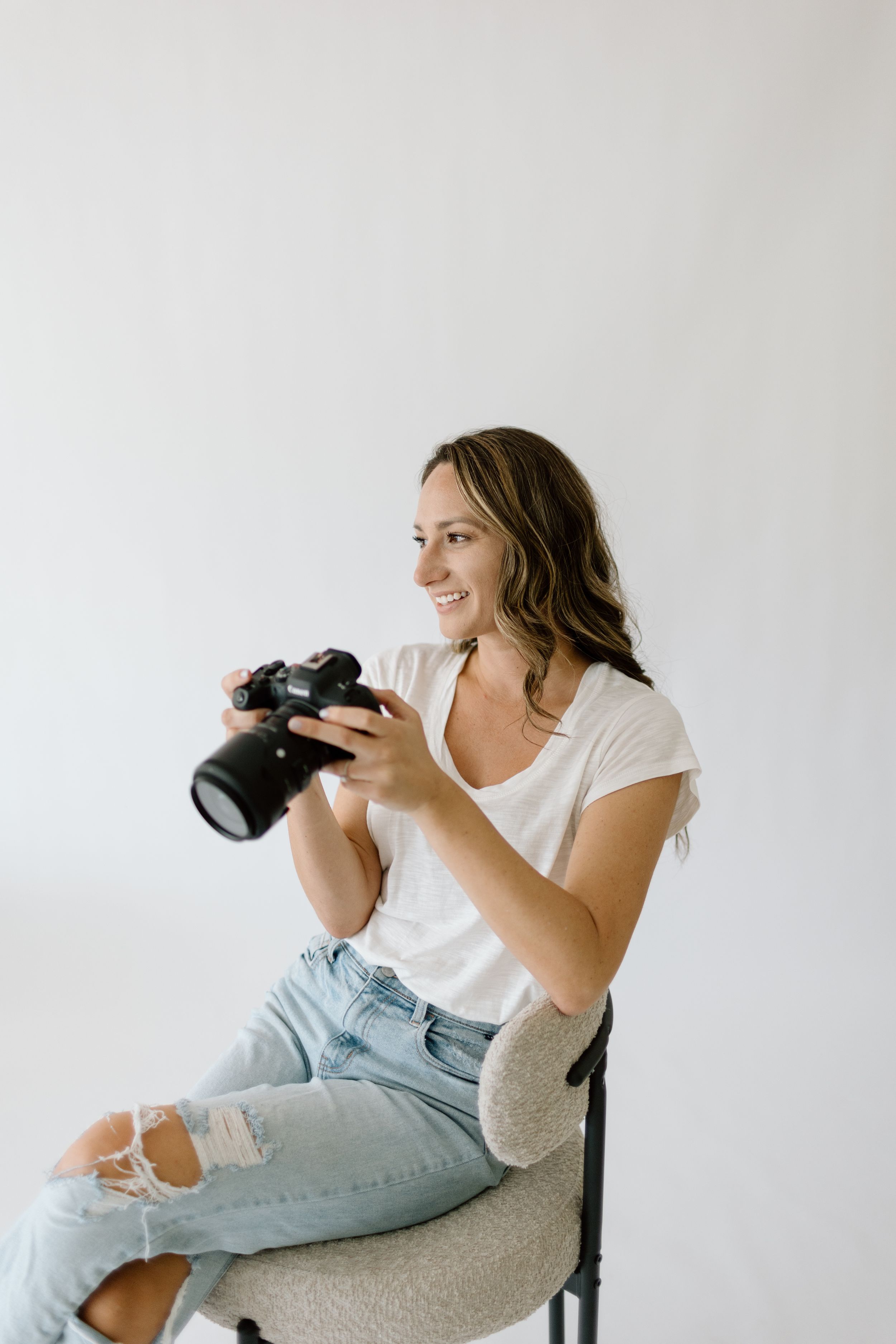 Photographer in casual white t-shirt and ripped jeans reviews images on professional camera while sitting against white wall.