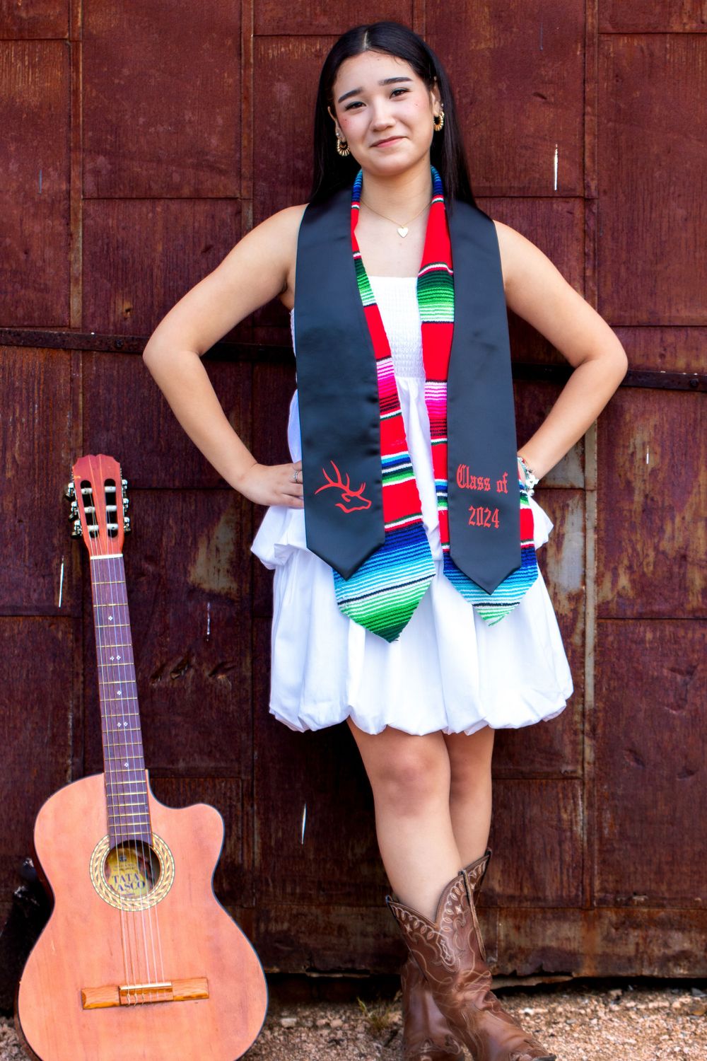 Graduate in white dress with decorative stole poses with acoustic guitar against rustic backdrop.