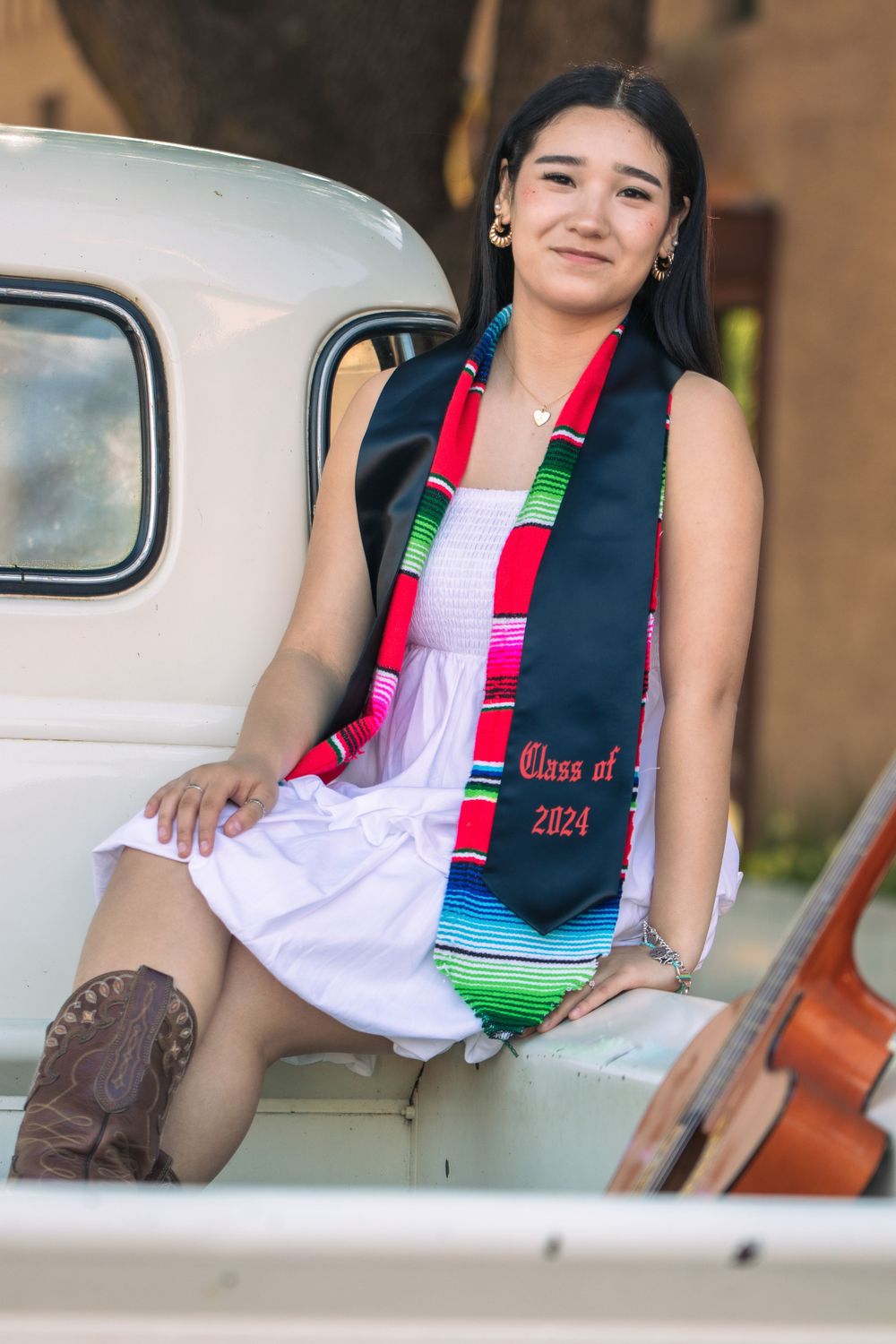 Graduate in white dress and stole poses with vintage car in historic setting.