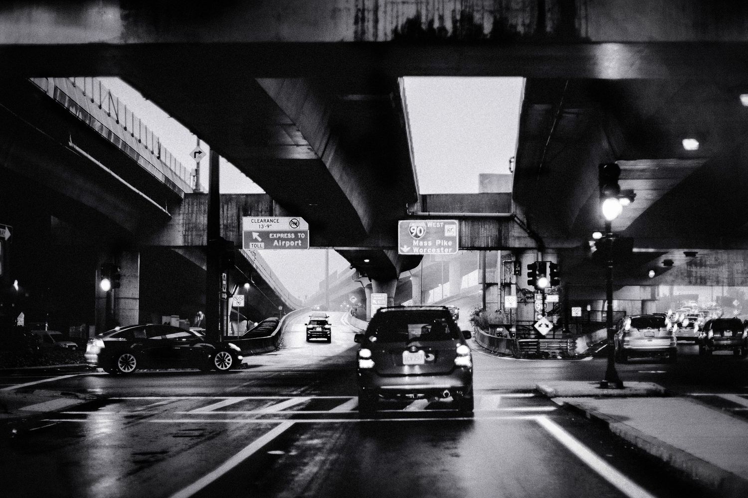 A black and white urban night scene showing cars under a dimly lit underpass with fluorescent lighting overhead.