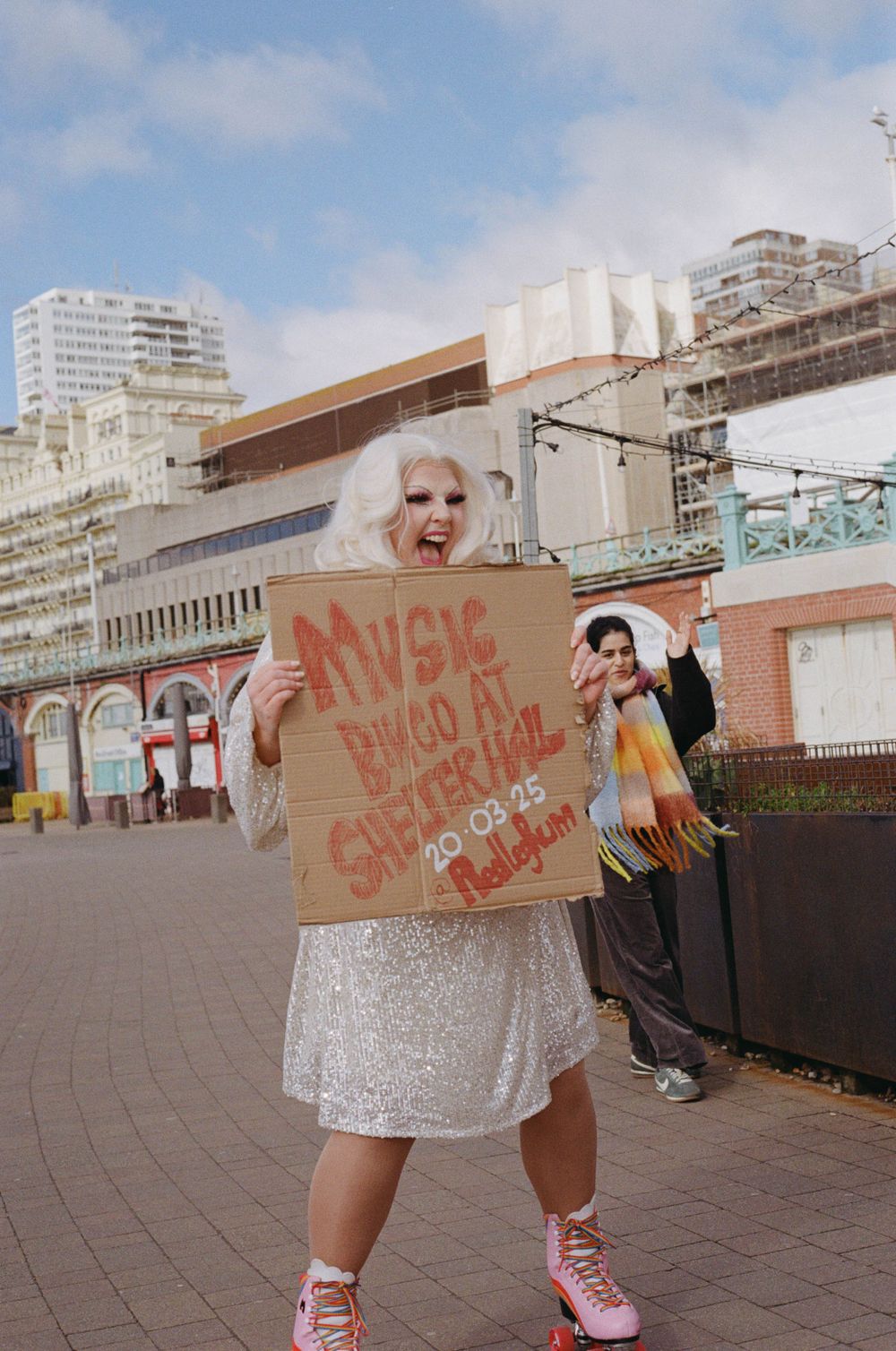 A person in a white dress holds a cardboard sign in front of an urban backdrop.