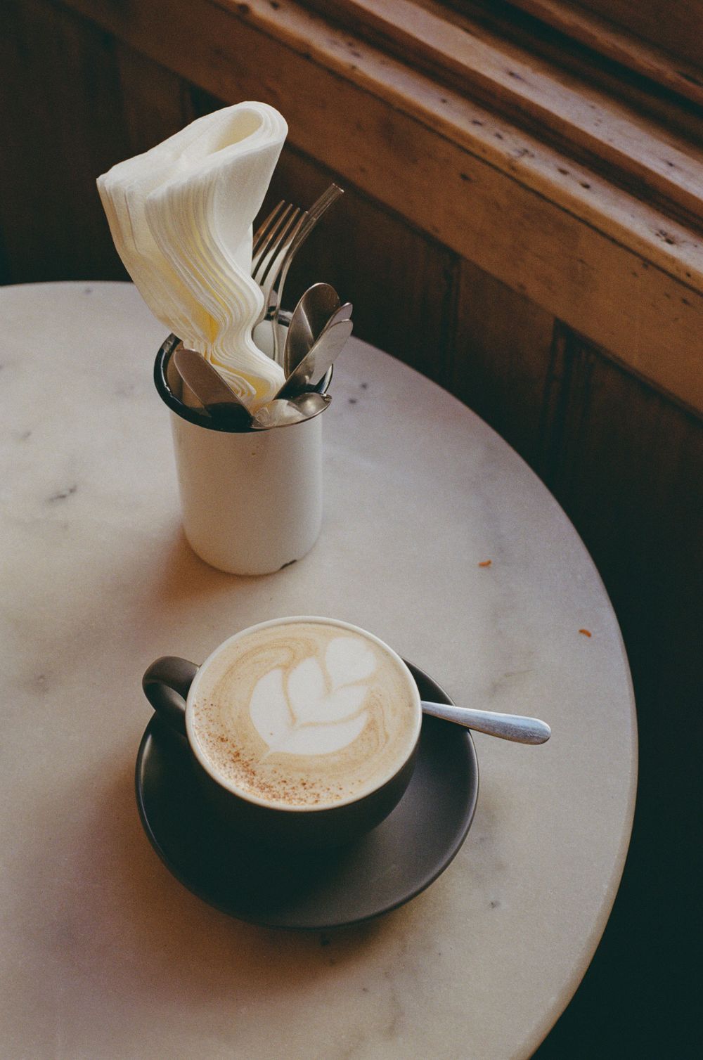 A cappuccino with latte art served alongside silverware in a white holder on a cafe table.