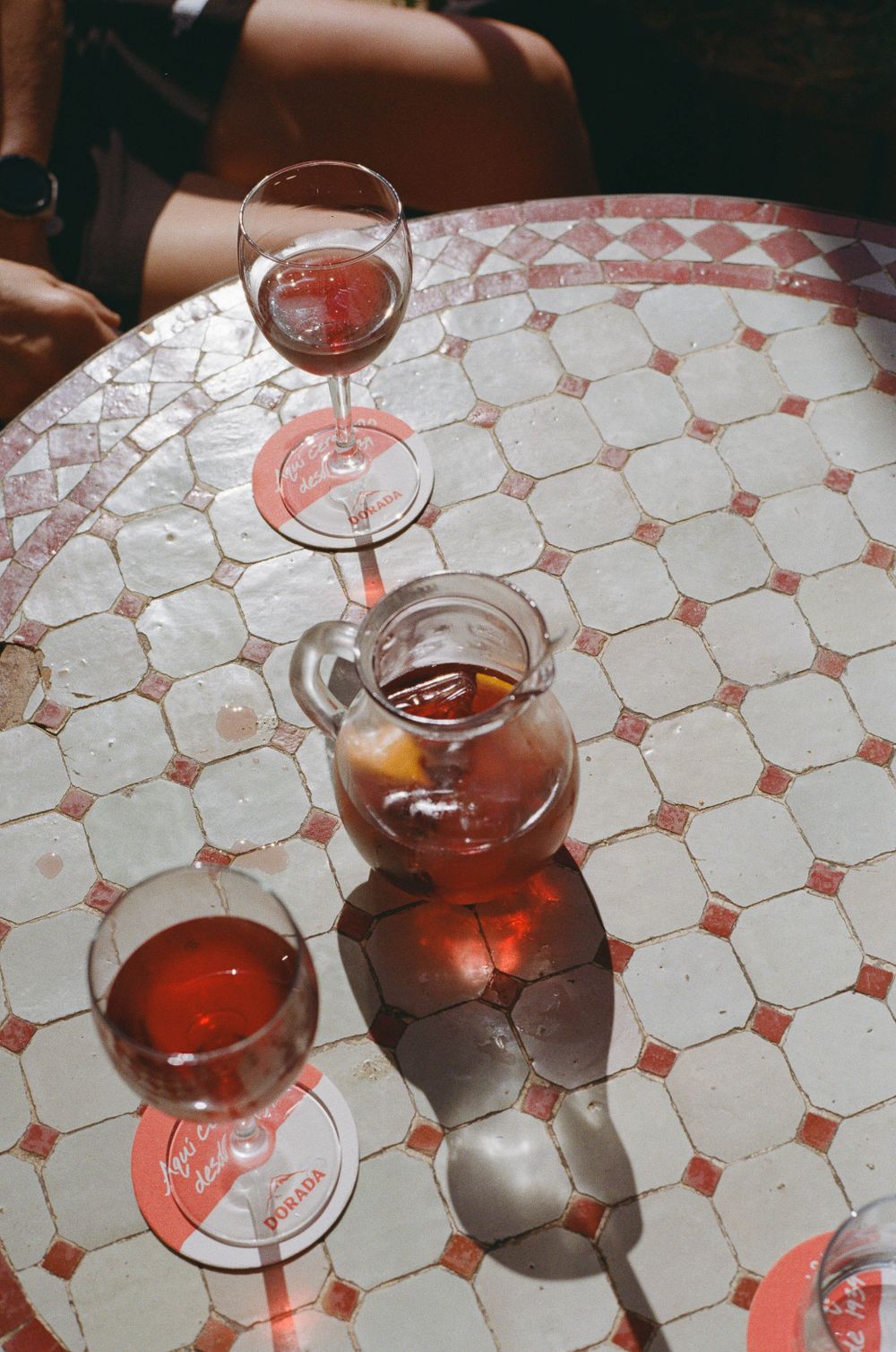 Rose wine glasses and carafe casting shadows on a patterned table surface in warm sunlight.
