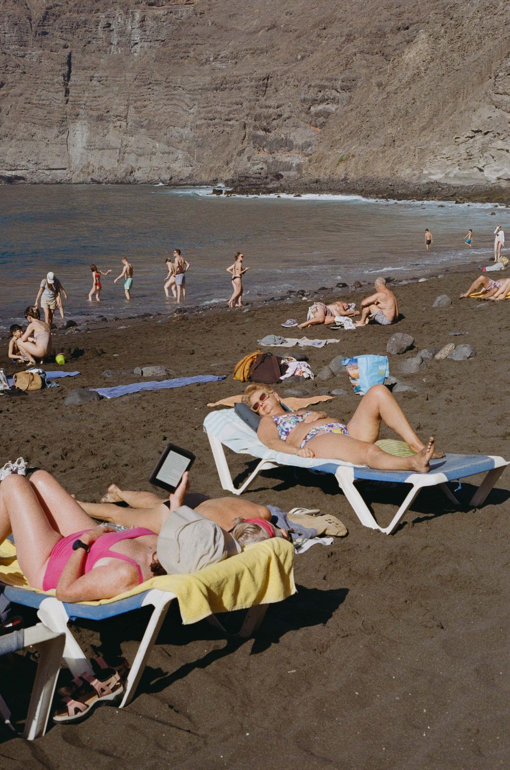 Beachgoers relaxing on lounge chairs on a dark sandy beach beneath towering cliffs.