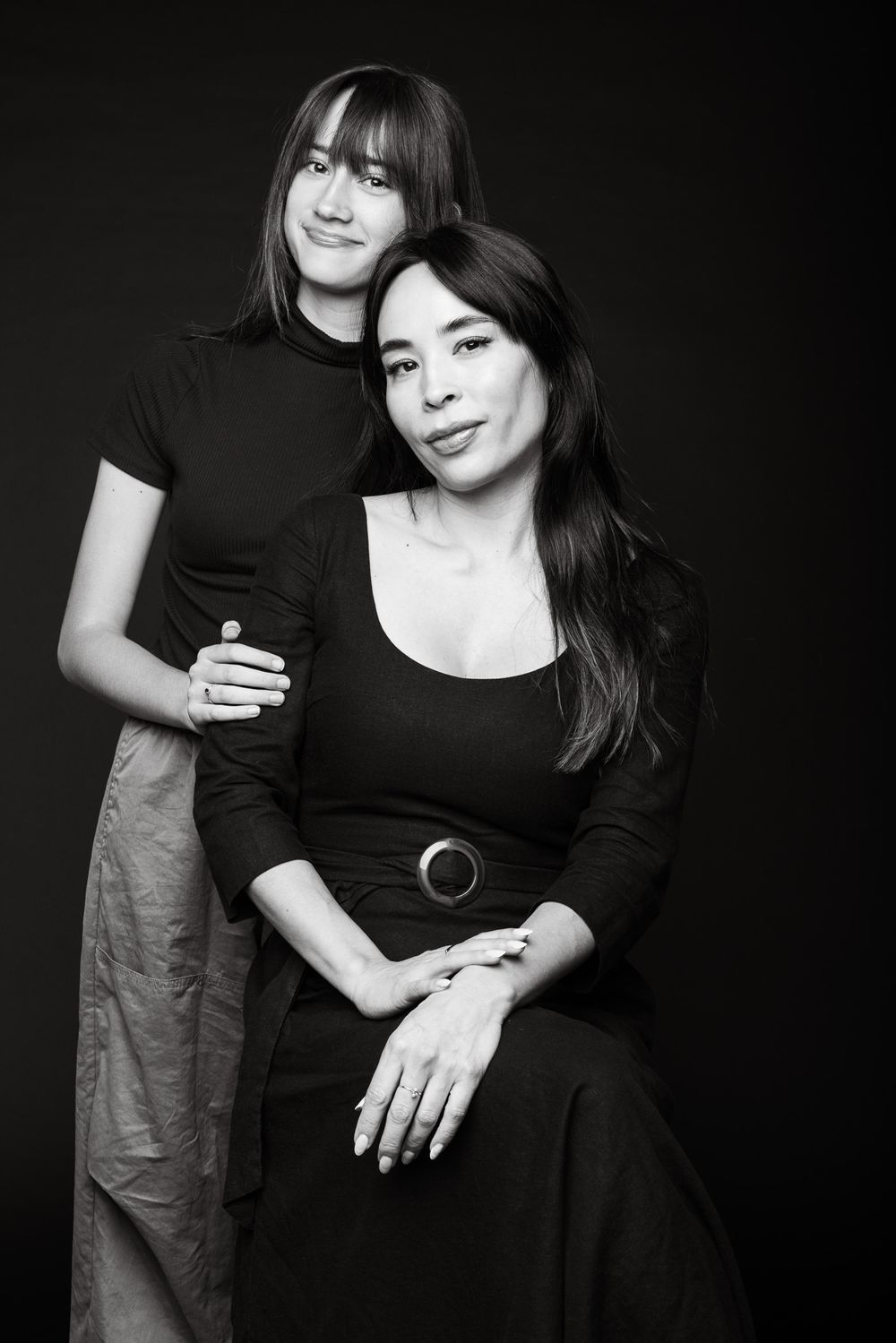 Two women in black outfits pose together for an artistic black and white portrait.