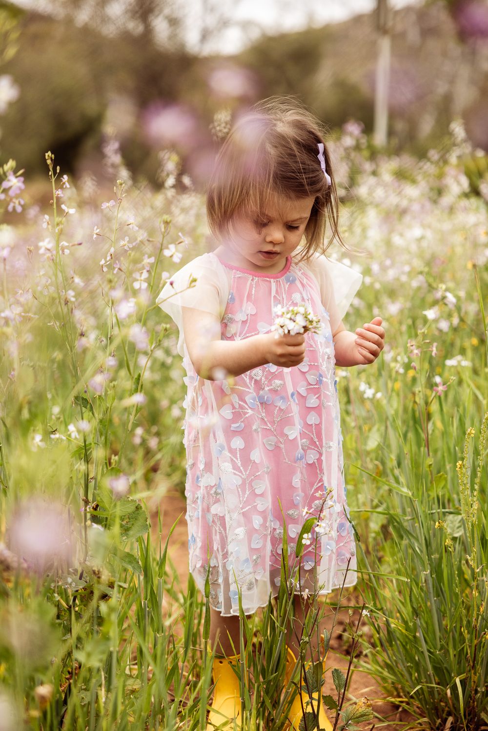 Child in a pink floral dress exploring wildflowers in a sunny meadow.
