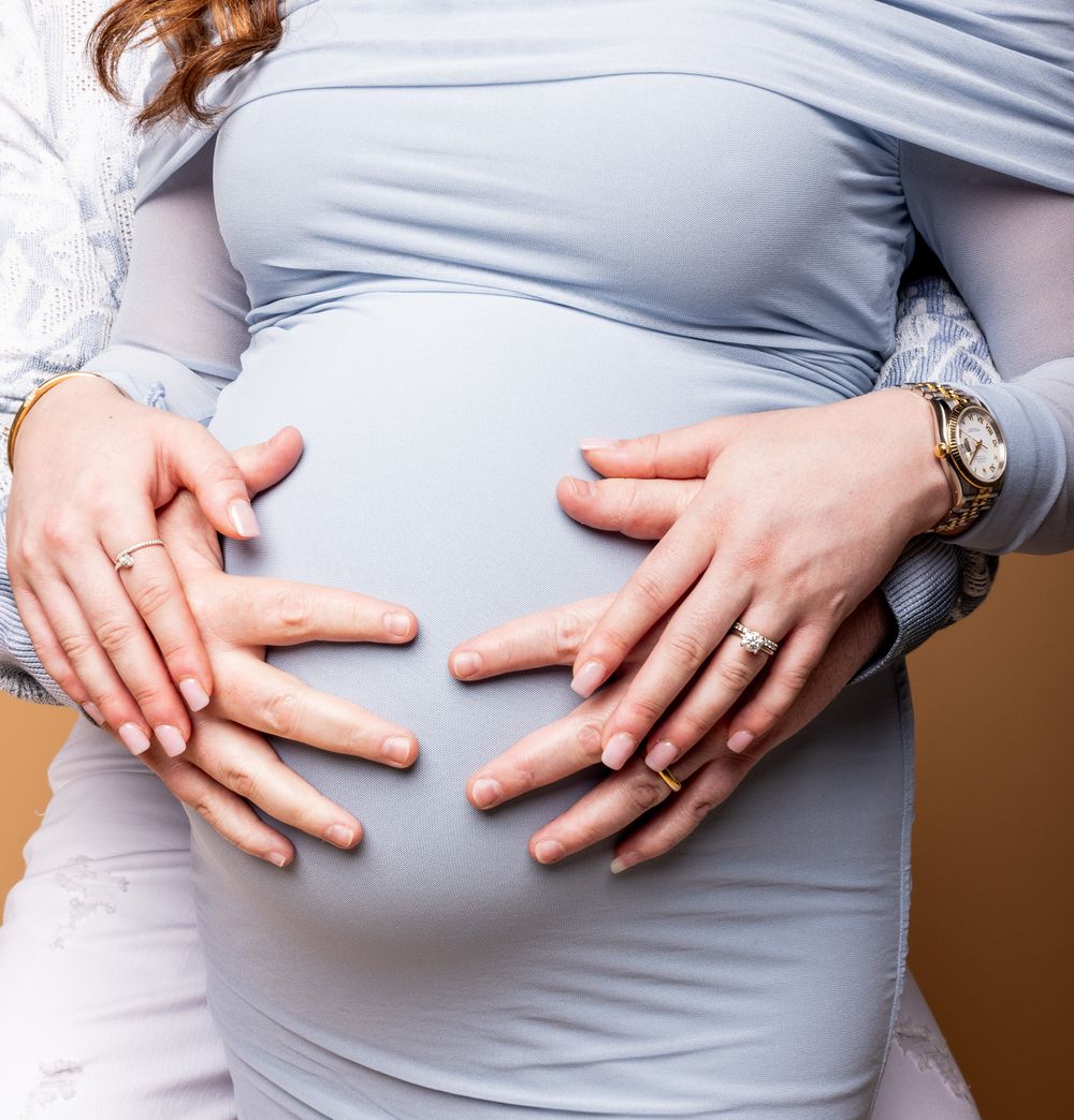 Close up of hands gently resting on a pregnant belly covered in light gray fabric.