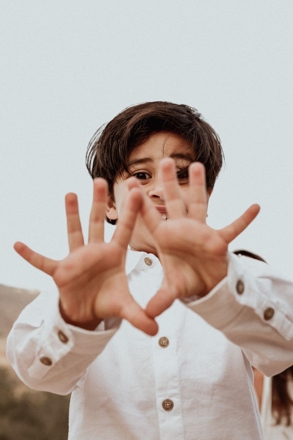 Child in white button-down shirt playfully holding hands up to camera.