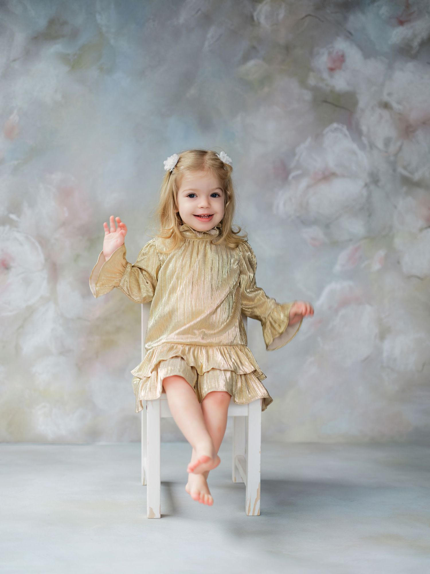 A small child wearing a frilly beige dress sits barefoot on a white wooden chair against a soft cloudy backdrop.