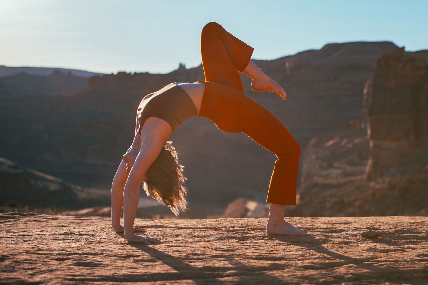 Silhouette of person doing backbend yoga pose against desert canyon backdrop at sunset.