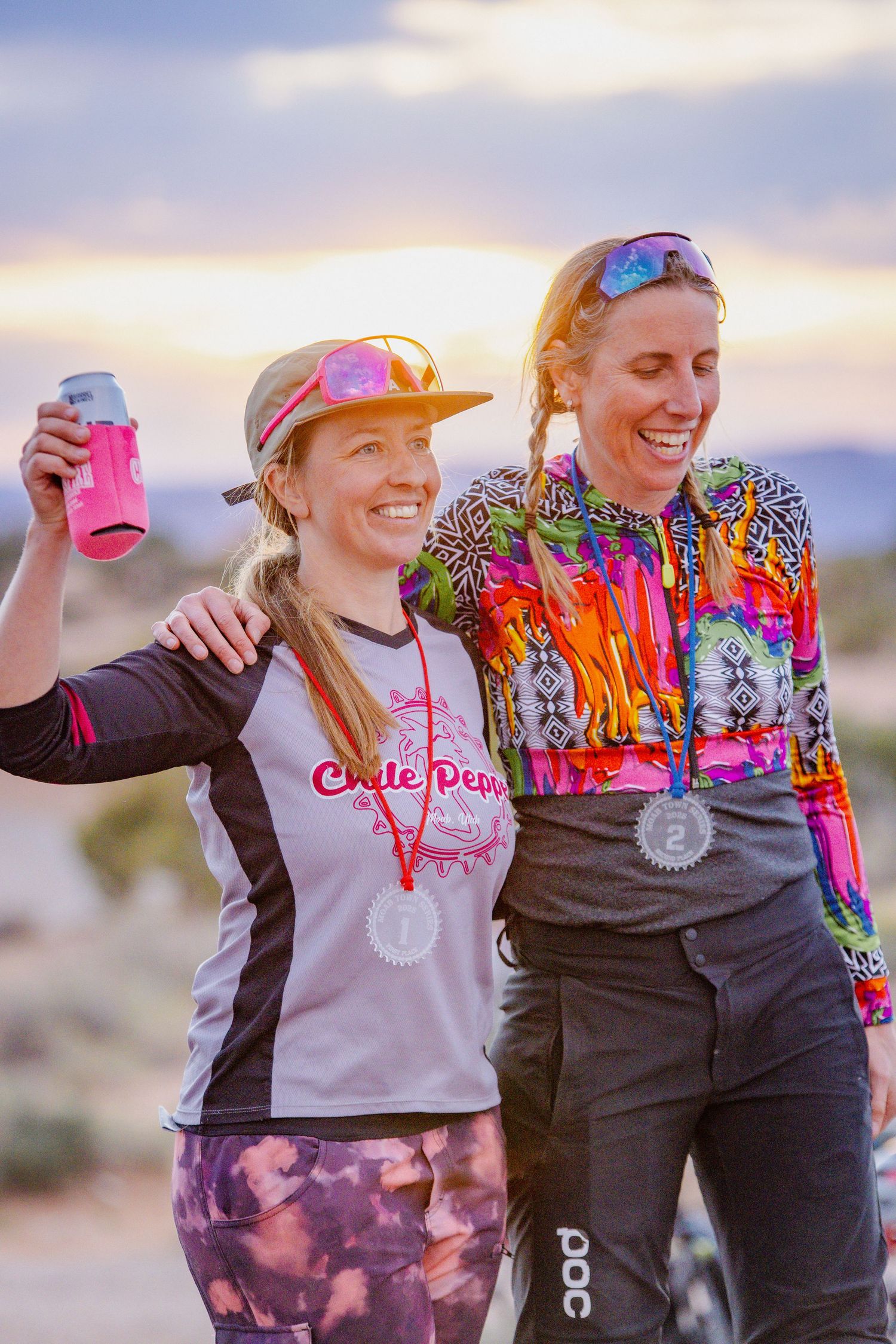Two athletes celebrating with drinks and medals after outdoor competition at sunset.