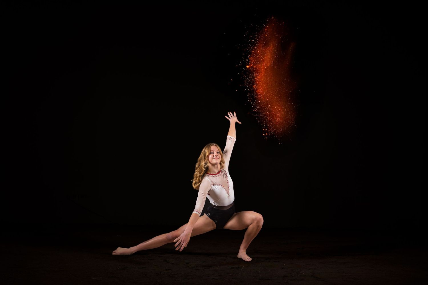 Dancer in black leotard performs dramatic pose with red powder effect against dark background.