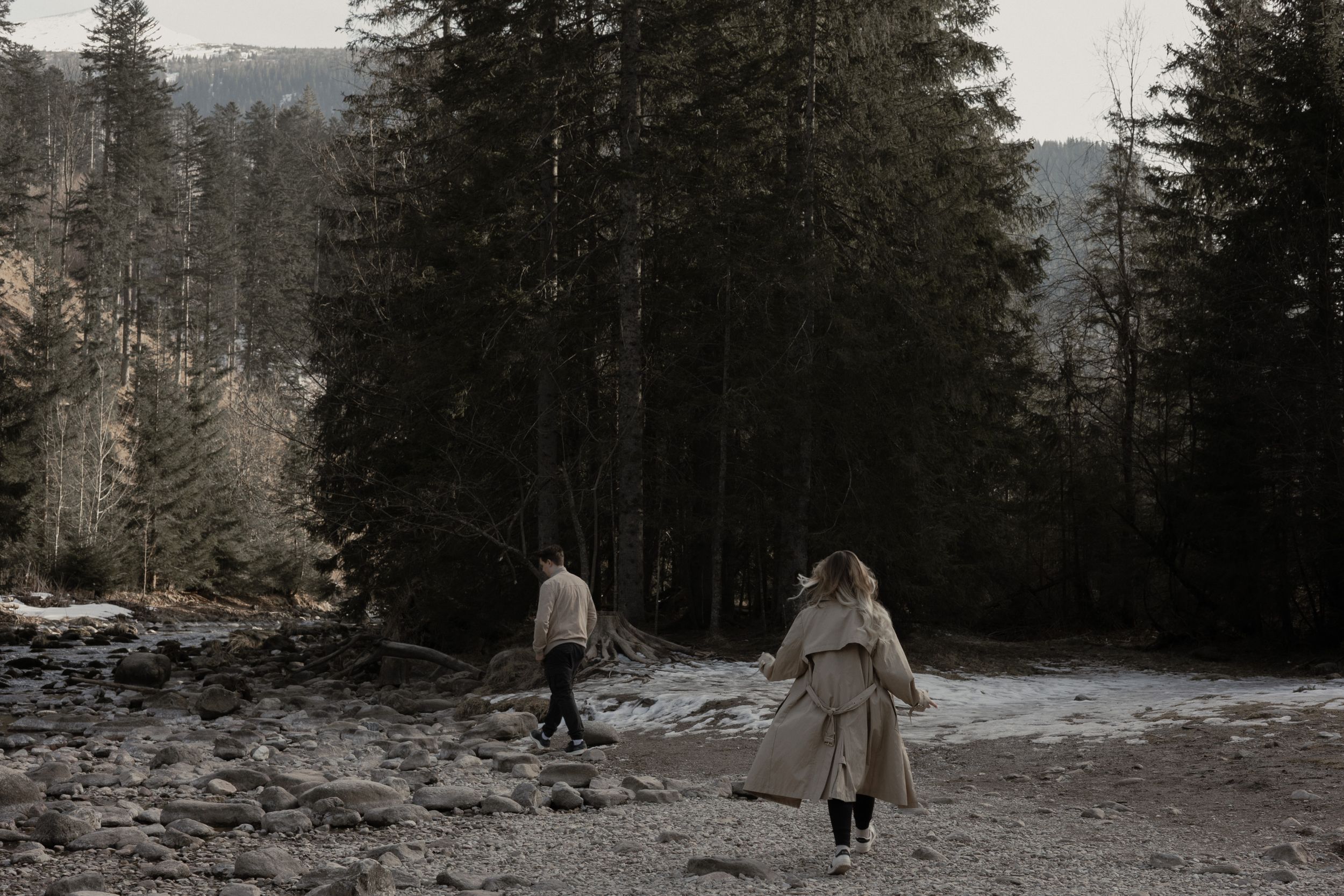 Two figures walk along a snowy forest path surrounded by tall evergreen trees on a winter day.