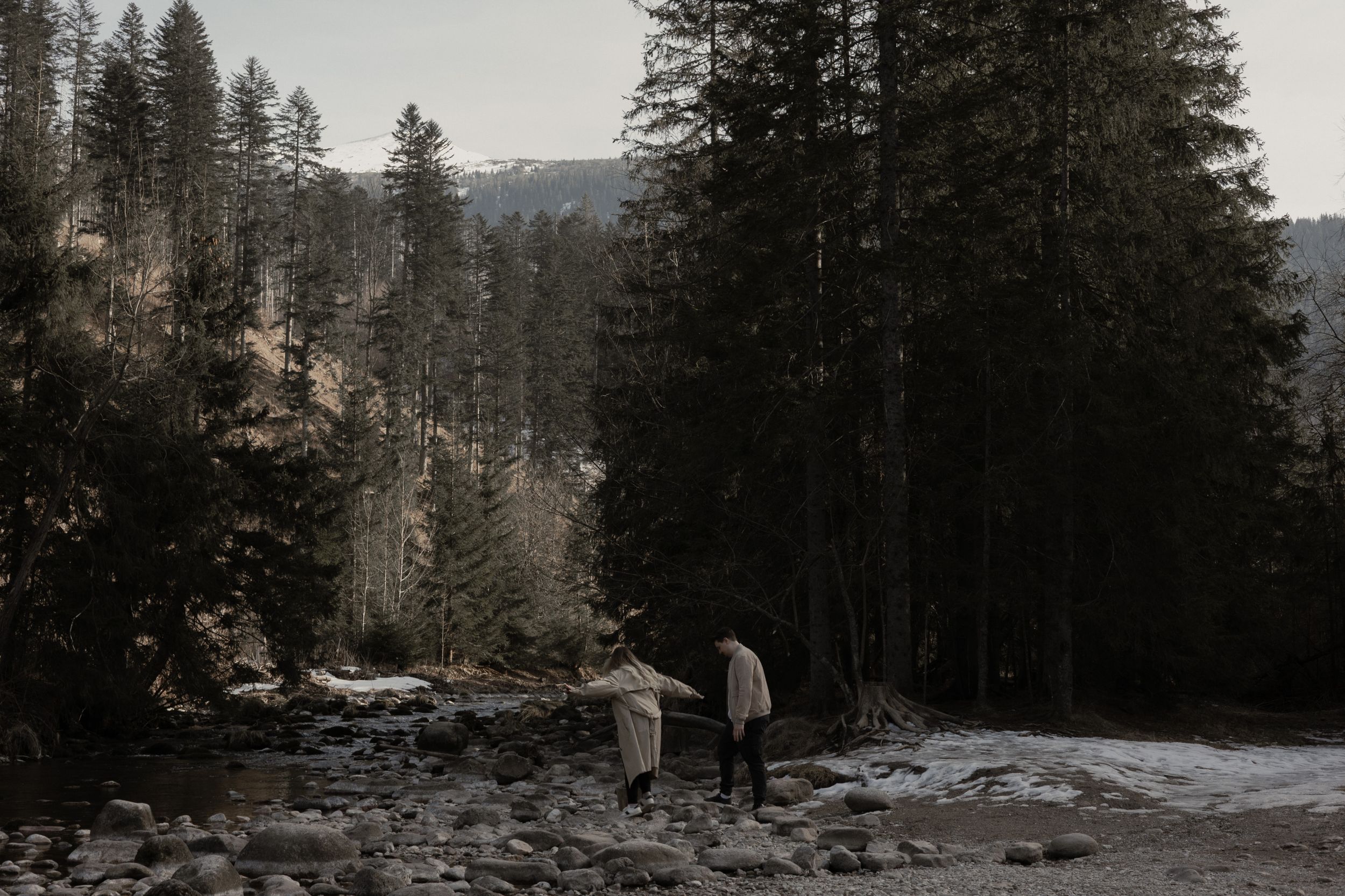 Two figures walk along a snowy mountain trail surrounded by towering evergreen trees in moody winter lighting.