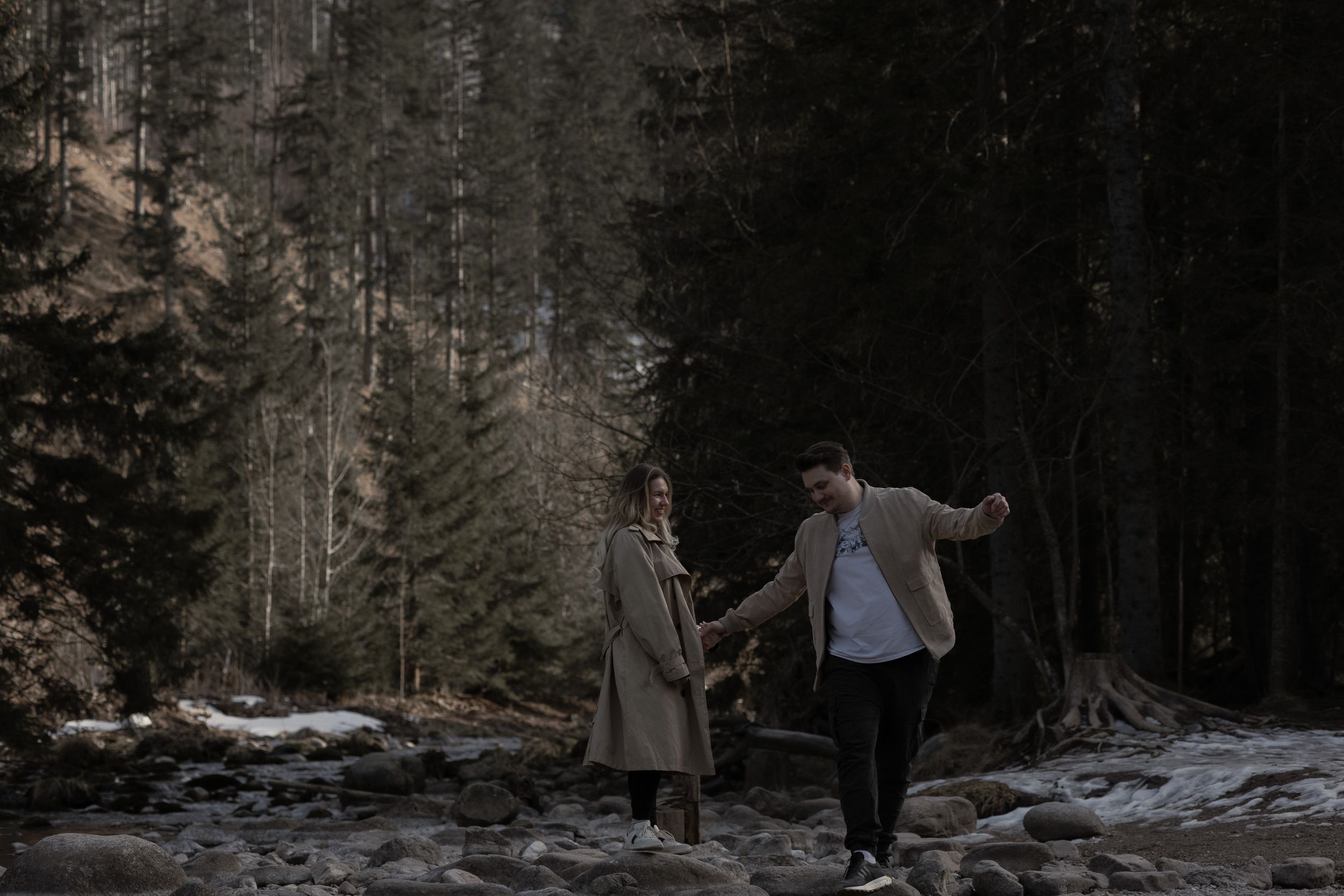 A couple holding hands walks together through a snowy winter forest path at dusk.