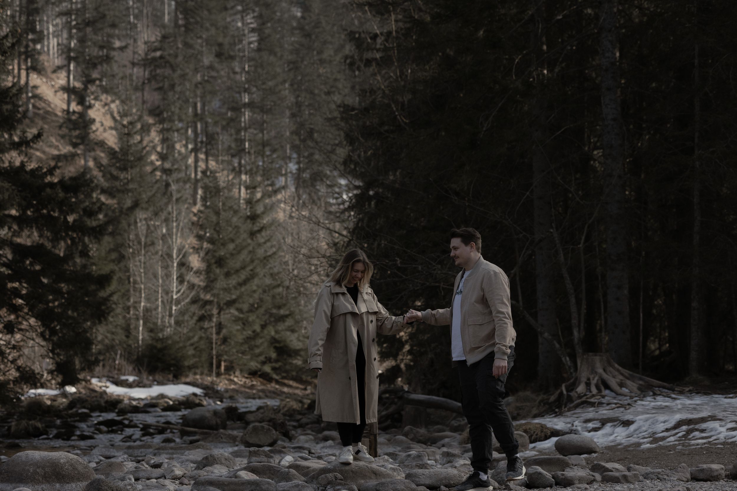 Two people stand among snowy evergreen trees in a dark moody winter forest setting.