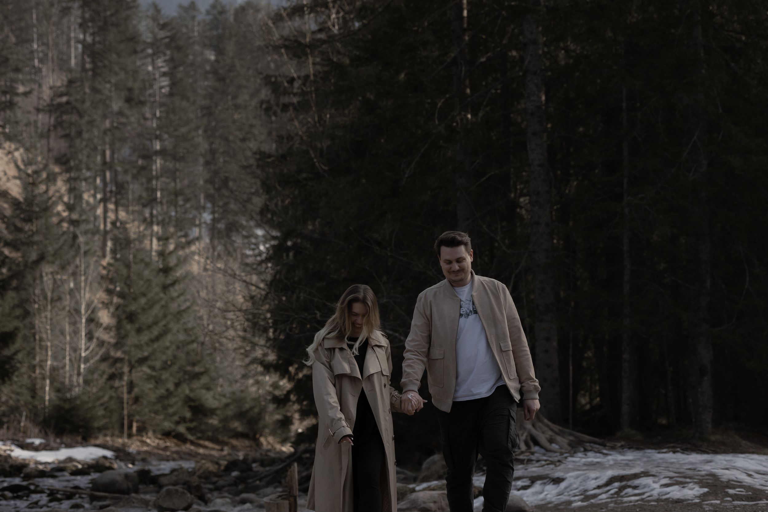A couple explores a wintry forest path with tall pine trees and patches of snow on the ground.