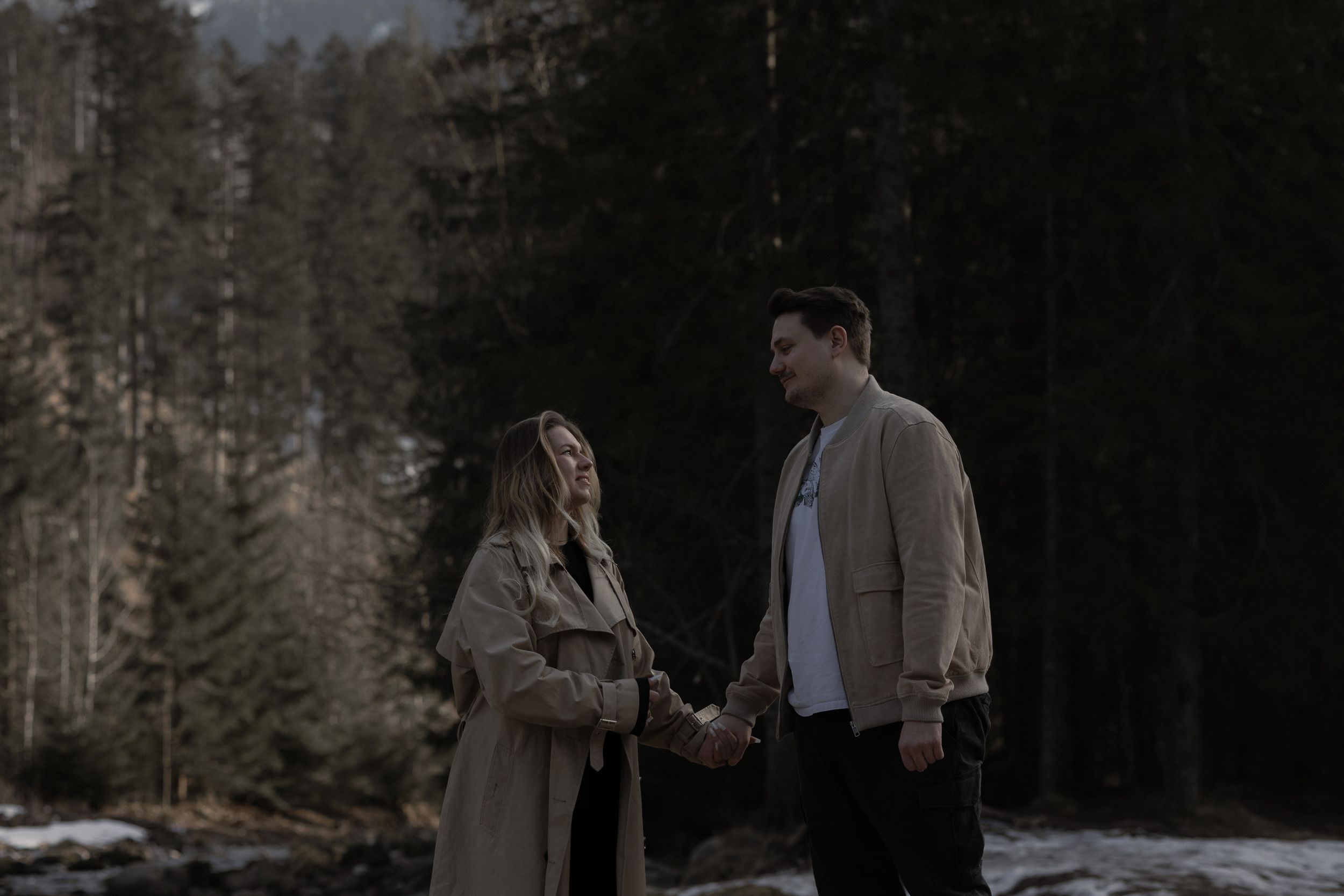 A couple stands holding hands in a moody forest setting at dusk wearing warm winter coats.