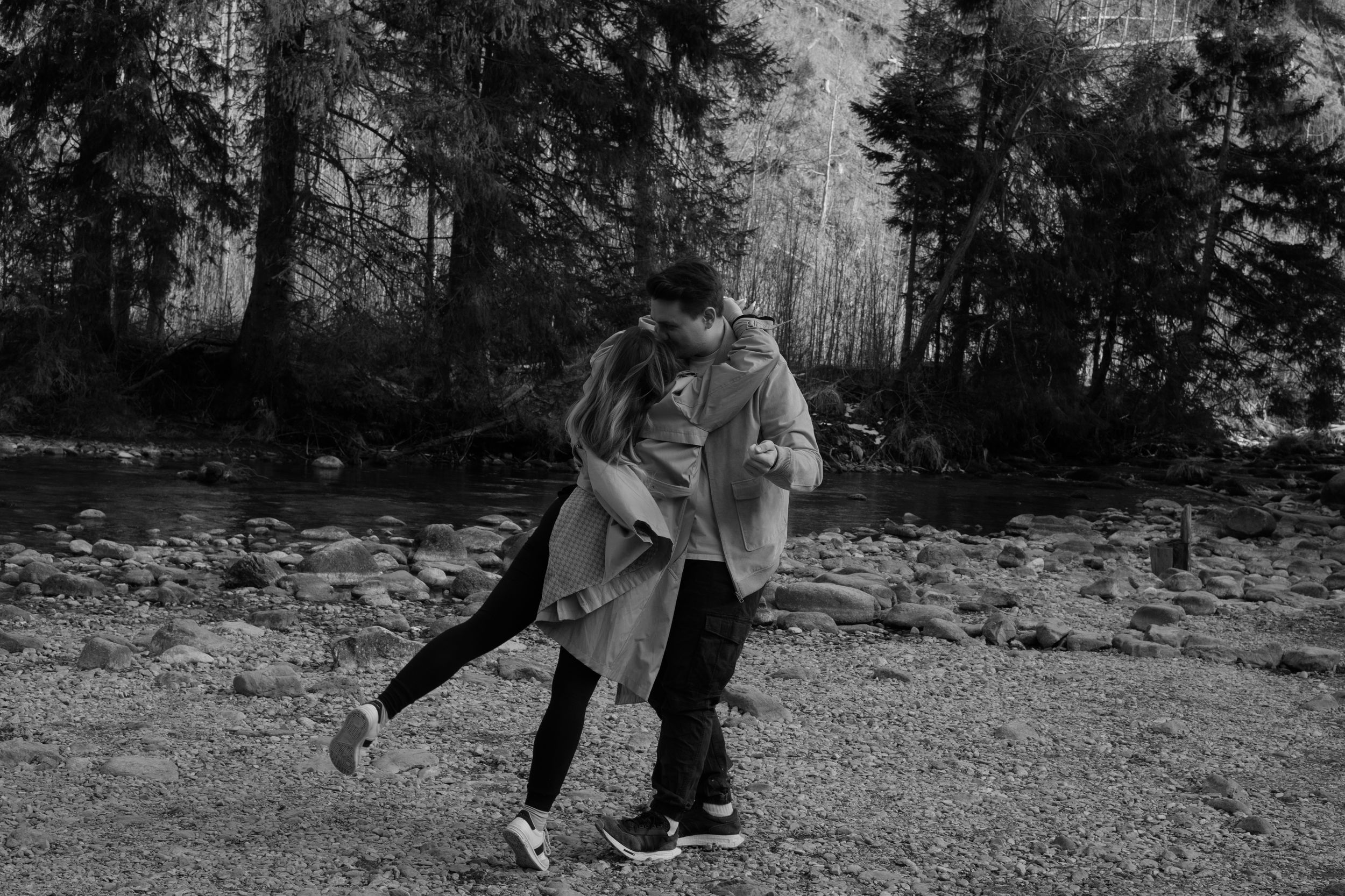 A couple shares a romantic embrace while dancing outdoors in a black and white photo near a forest setting.