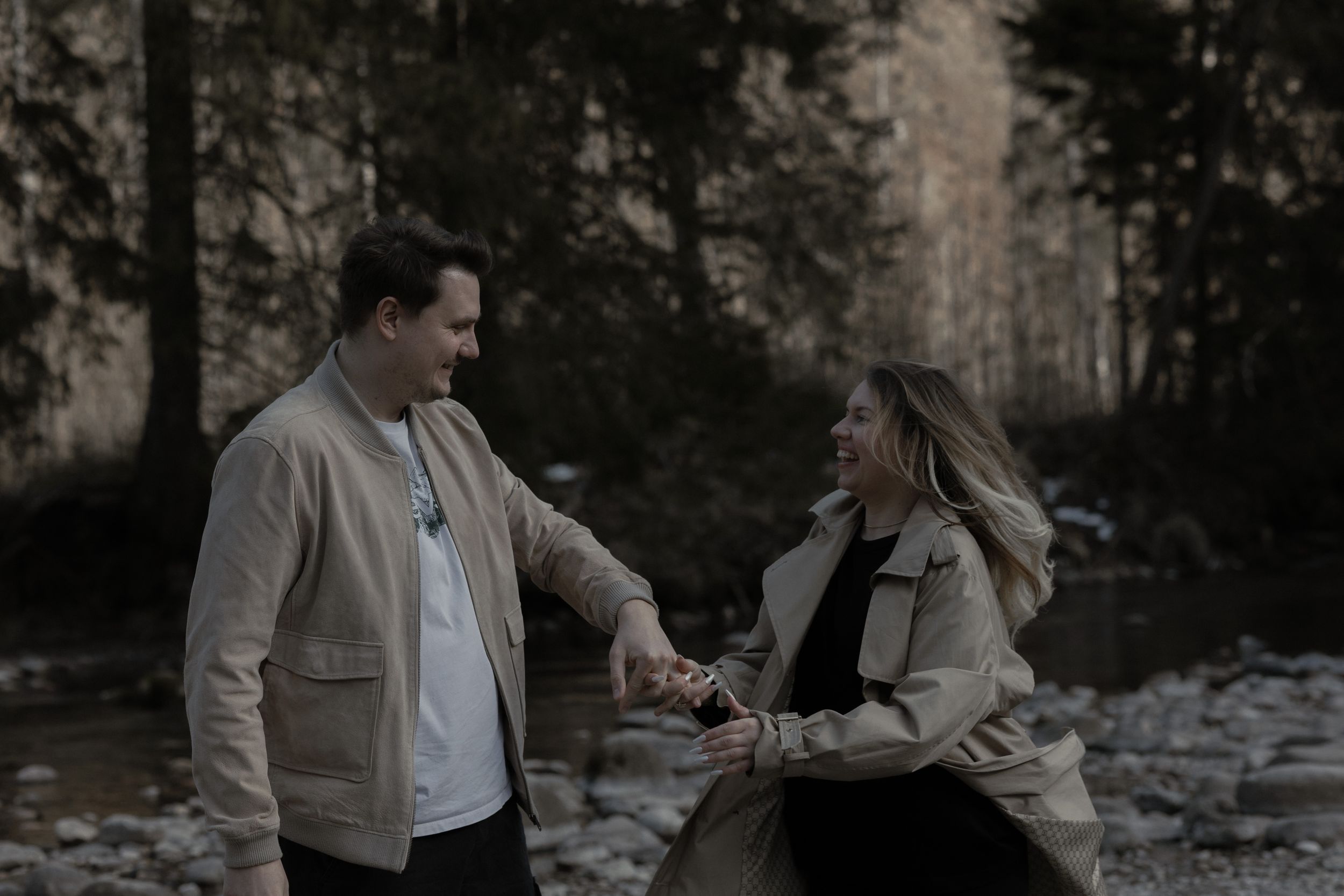 A couple holding hands and sharing a playful moment in a moody forest setting beside a rocky stream.