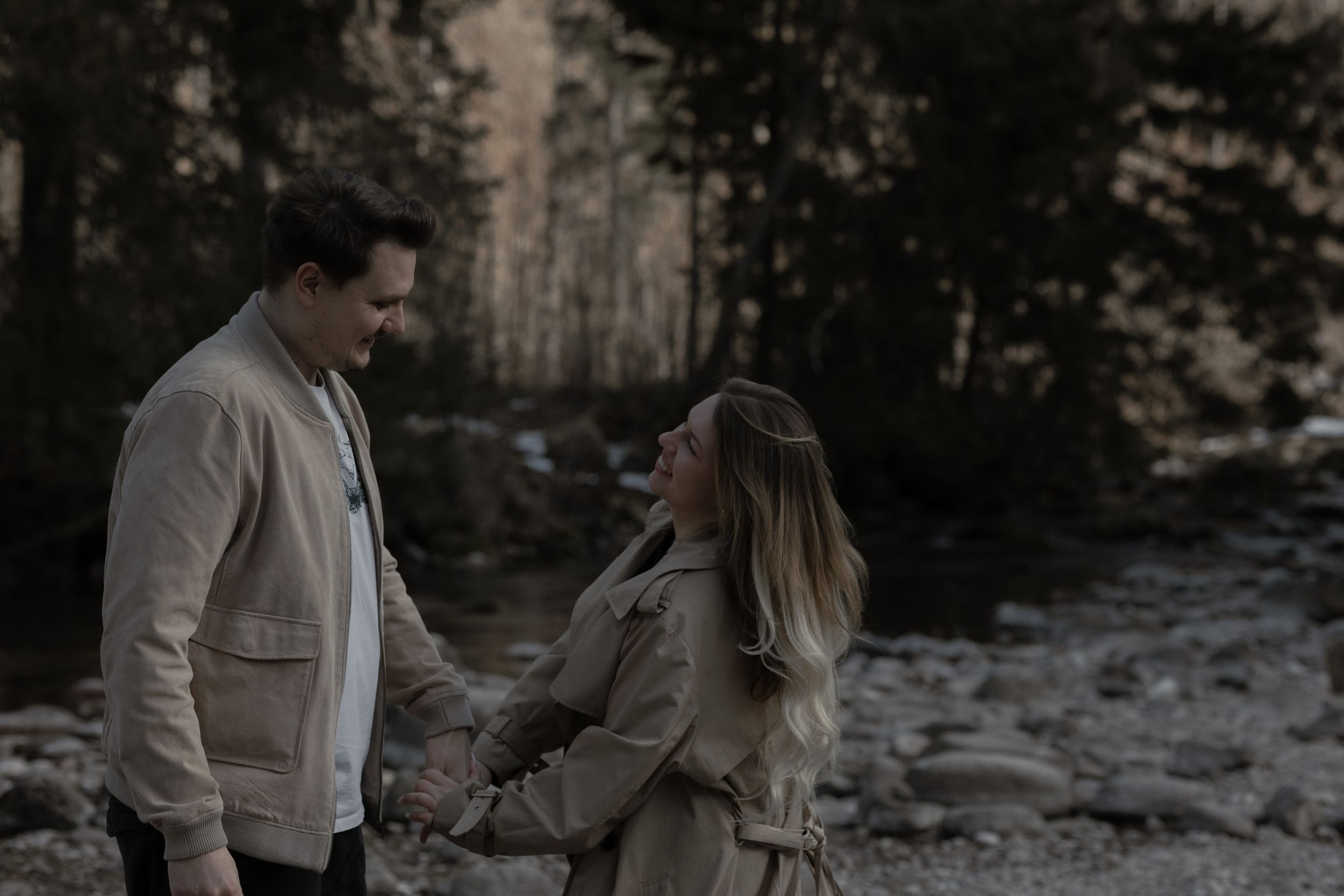 A couple in warm coats holds hands while standing near a snowy forest landscape during winter.