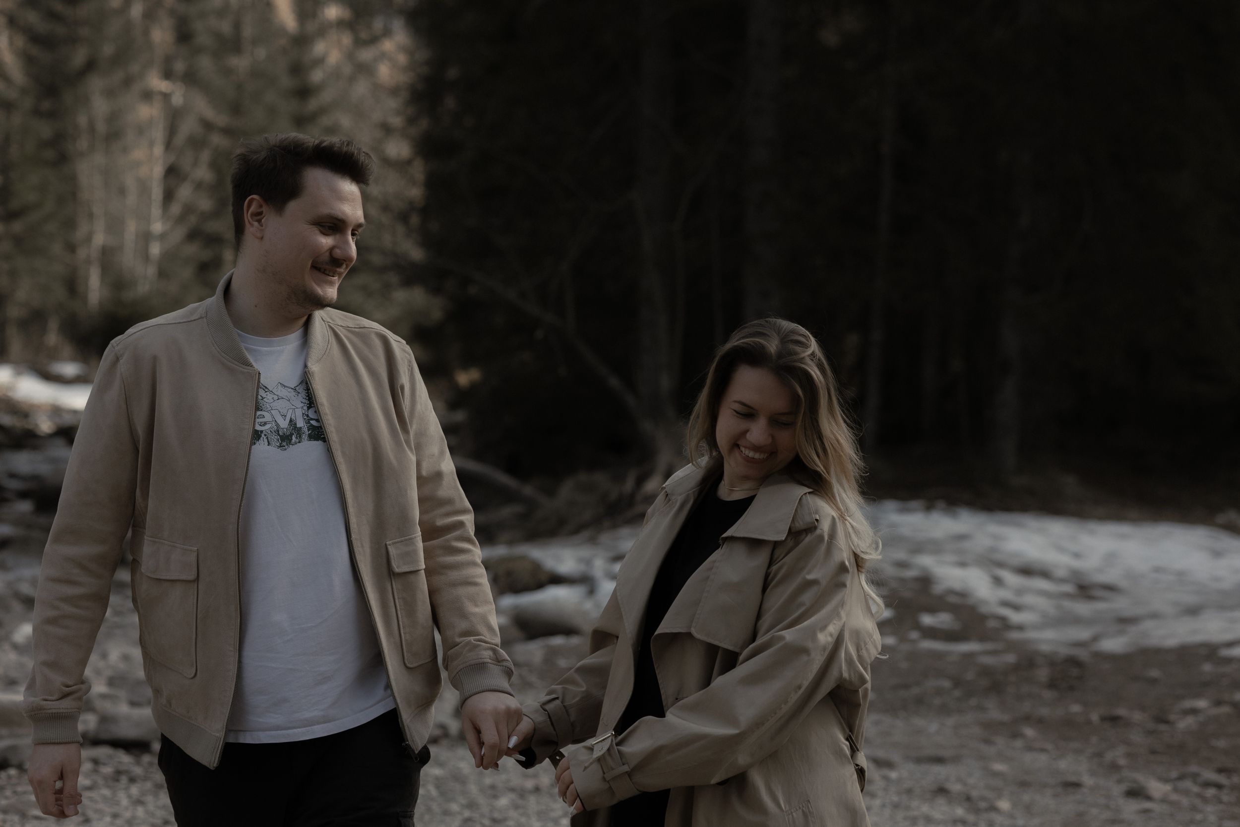 A couple walks together along a rocky creek bed in winter wearing light jackets and sharing a candid moment.