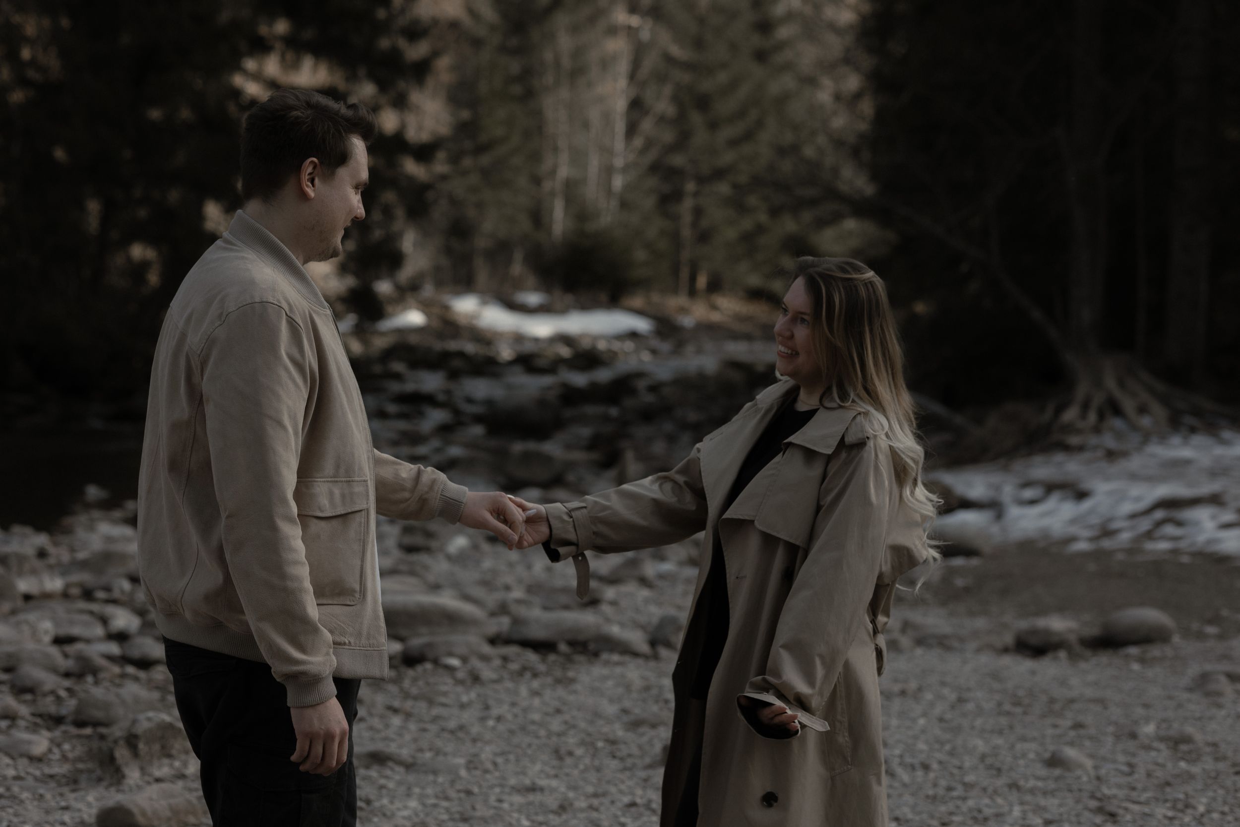 Two people in coats stand facing each other on a snowy riverbank in a moody winter forest setting.