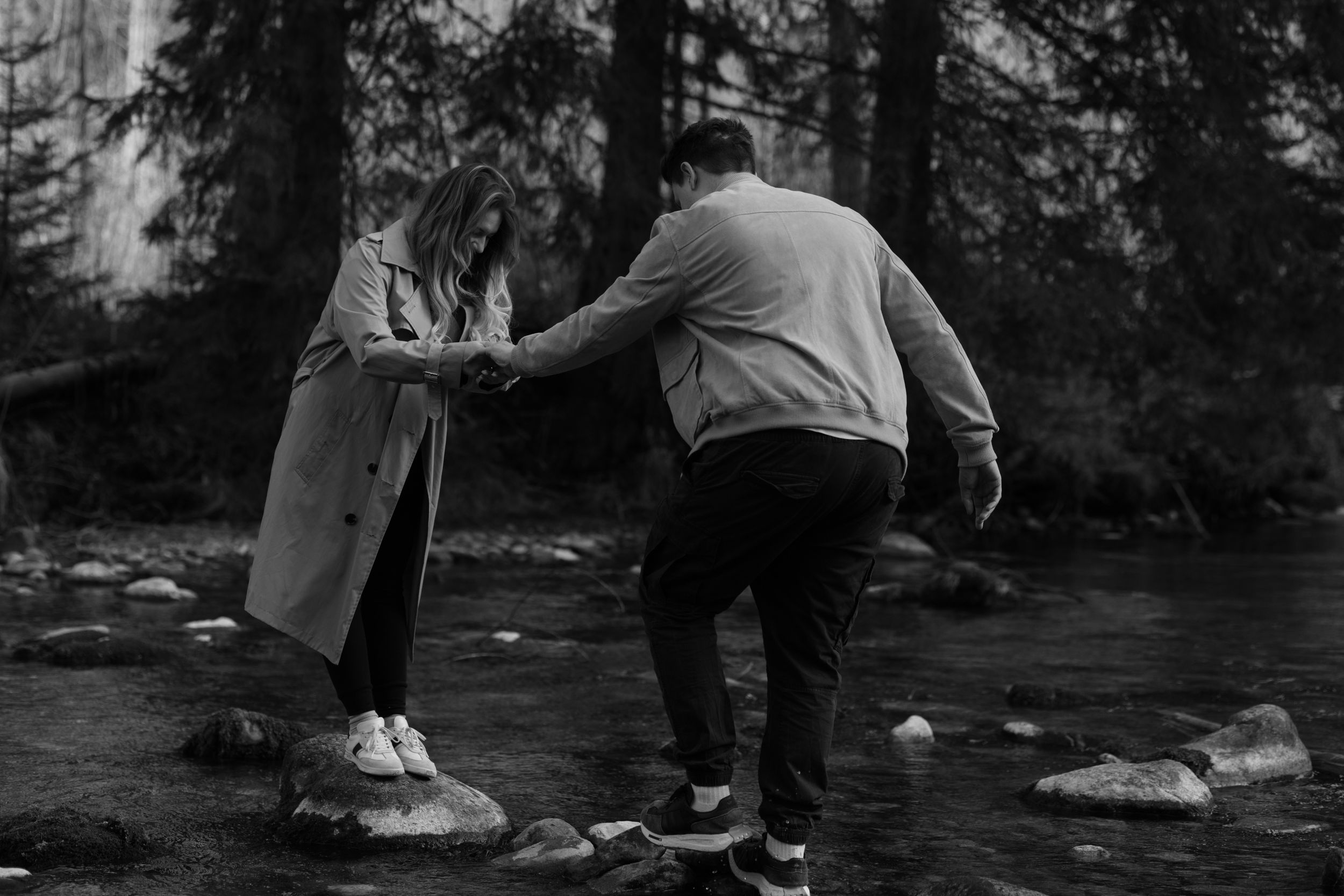 Two people balancing and walking on rocks along a misty creek in a forest setting.