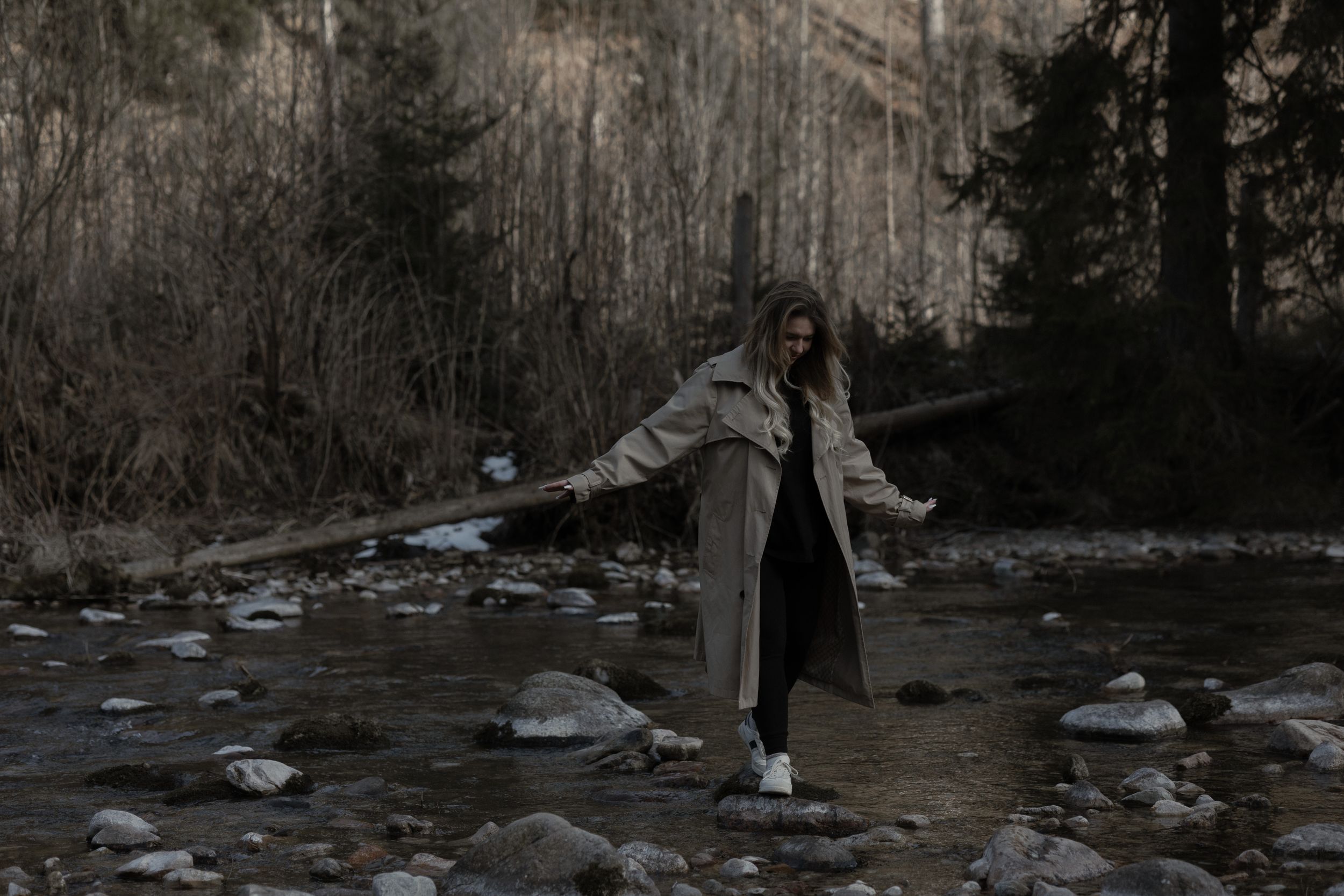 Person in a long coat balances with outstretched arms on river rocks during a moody forest evening.