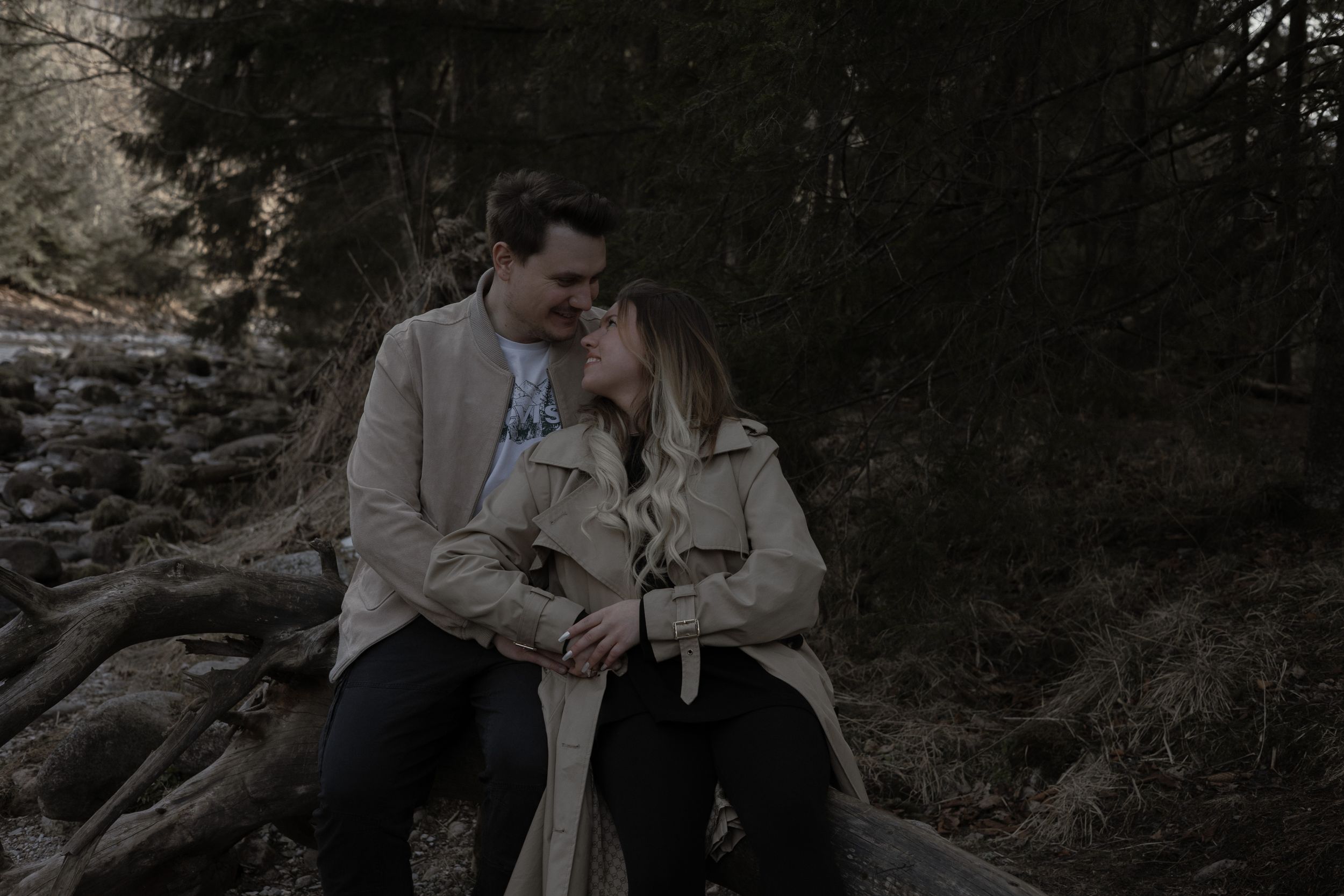 Couple in casual attire share an intimate moment while sitting on fallen logs in a forest setting.