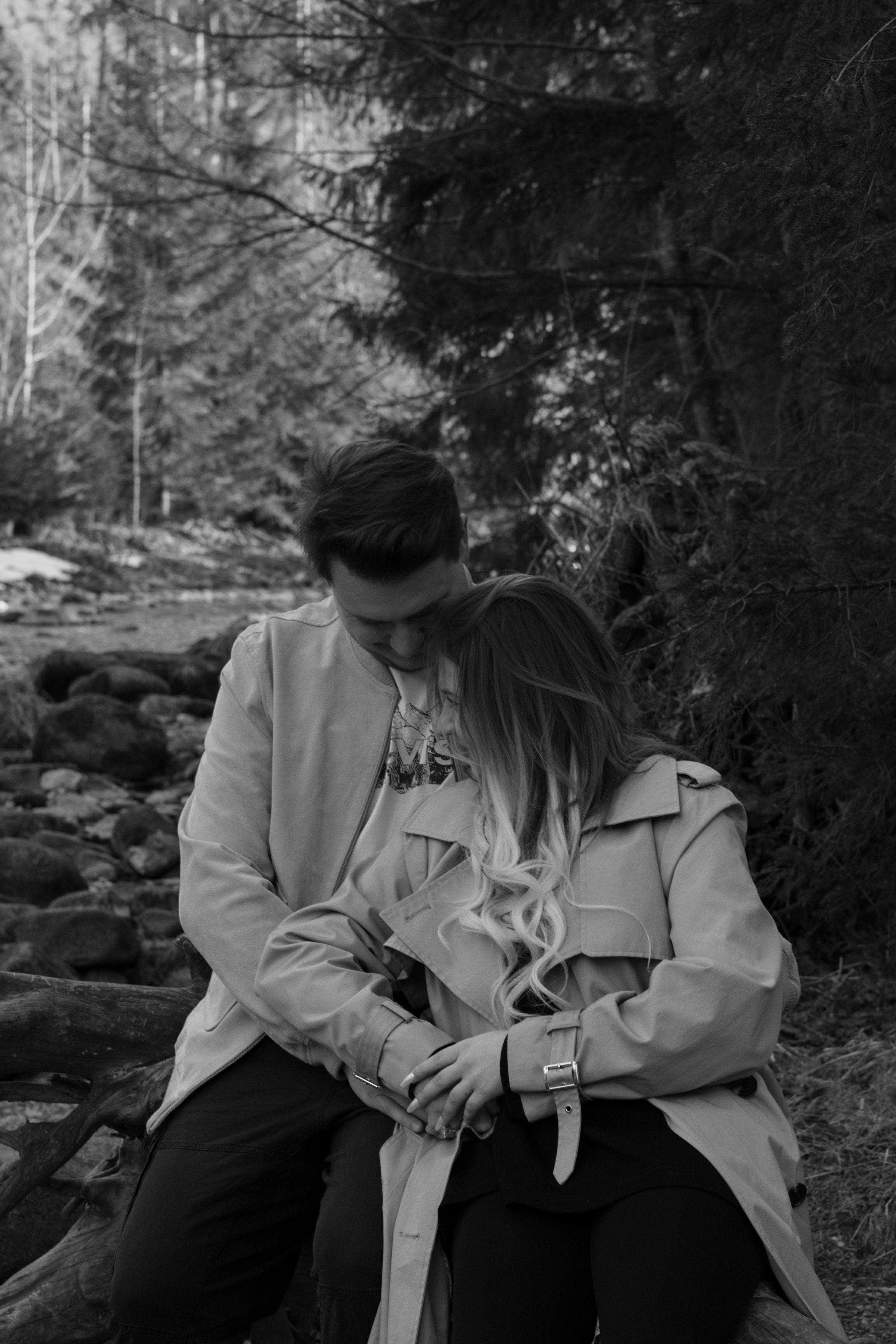 A couple in light colored jackets share an intimate moment together on rocks in a wooded forest setting.