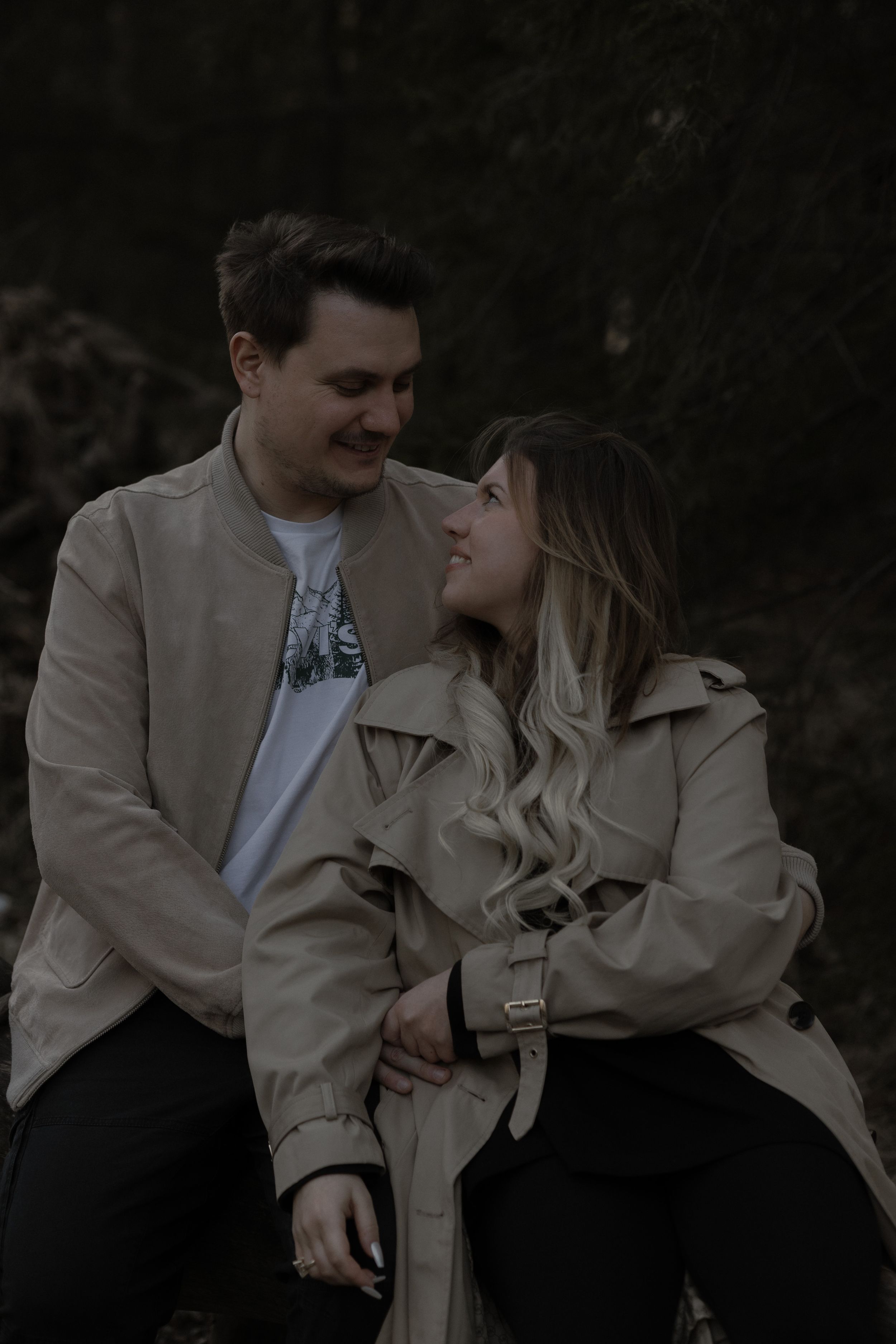 A romantic couple in beige coats sharing an intimate moment against a dark moody background.