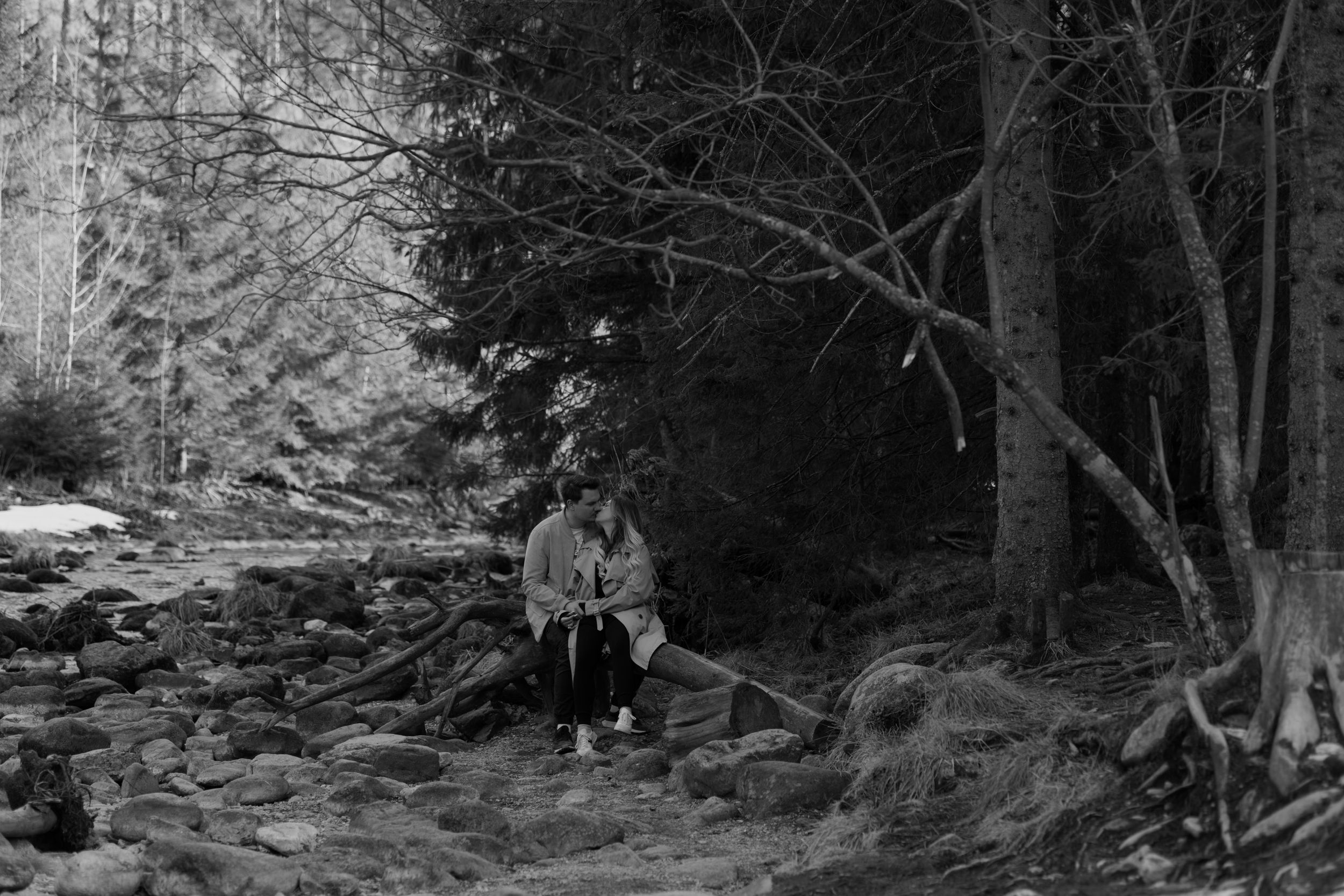 A black and white vintage photograph shows a person with a fishing pole sitting by a rocky forest stream.