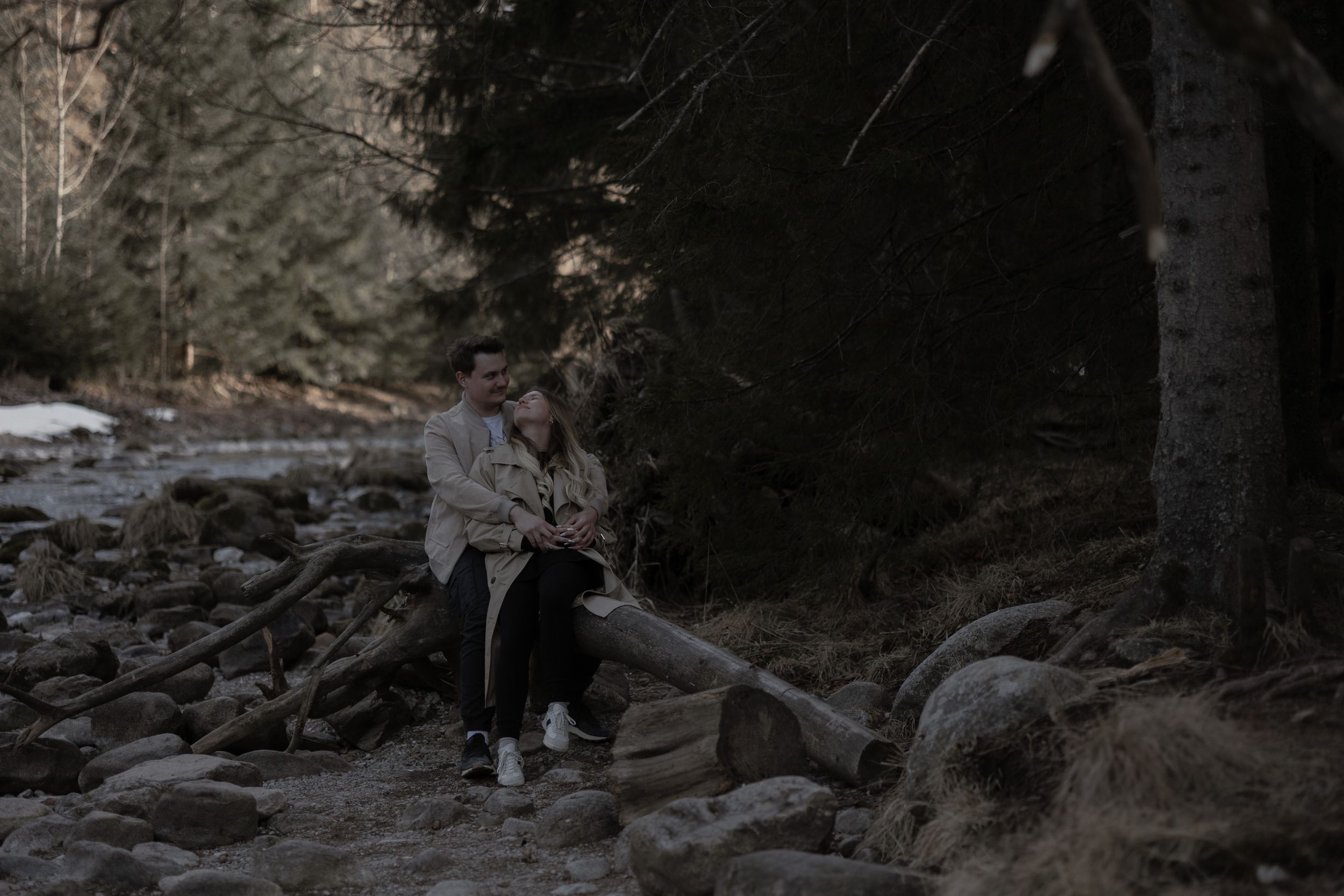 Figure sits on a fallen tree trunk in a moody forest setting with scattered snow and rocks on the ground.