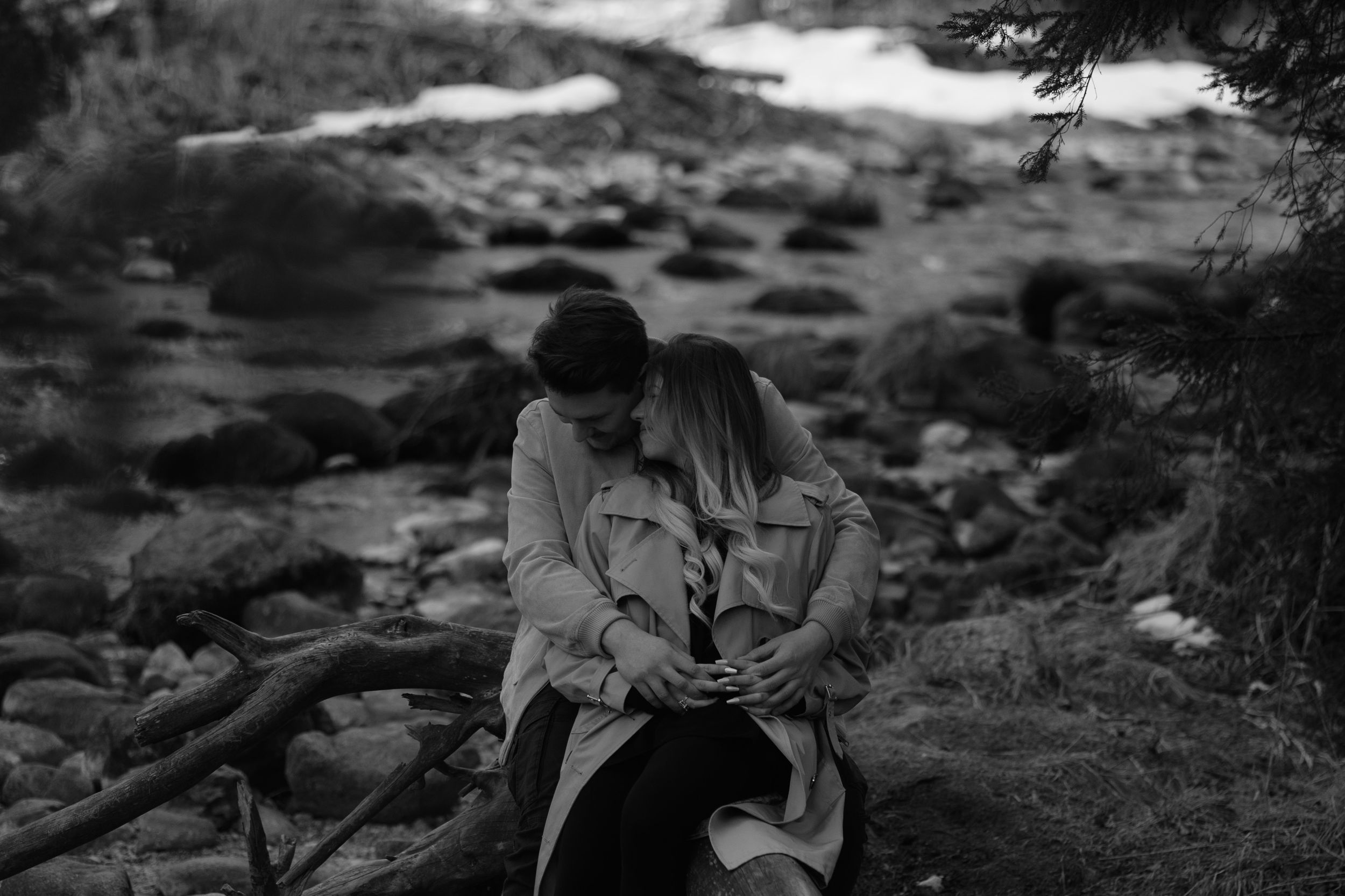 Black and white romantic scene of a couple embracing by a rocky stream in nature.
