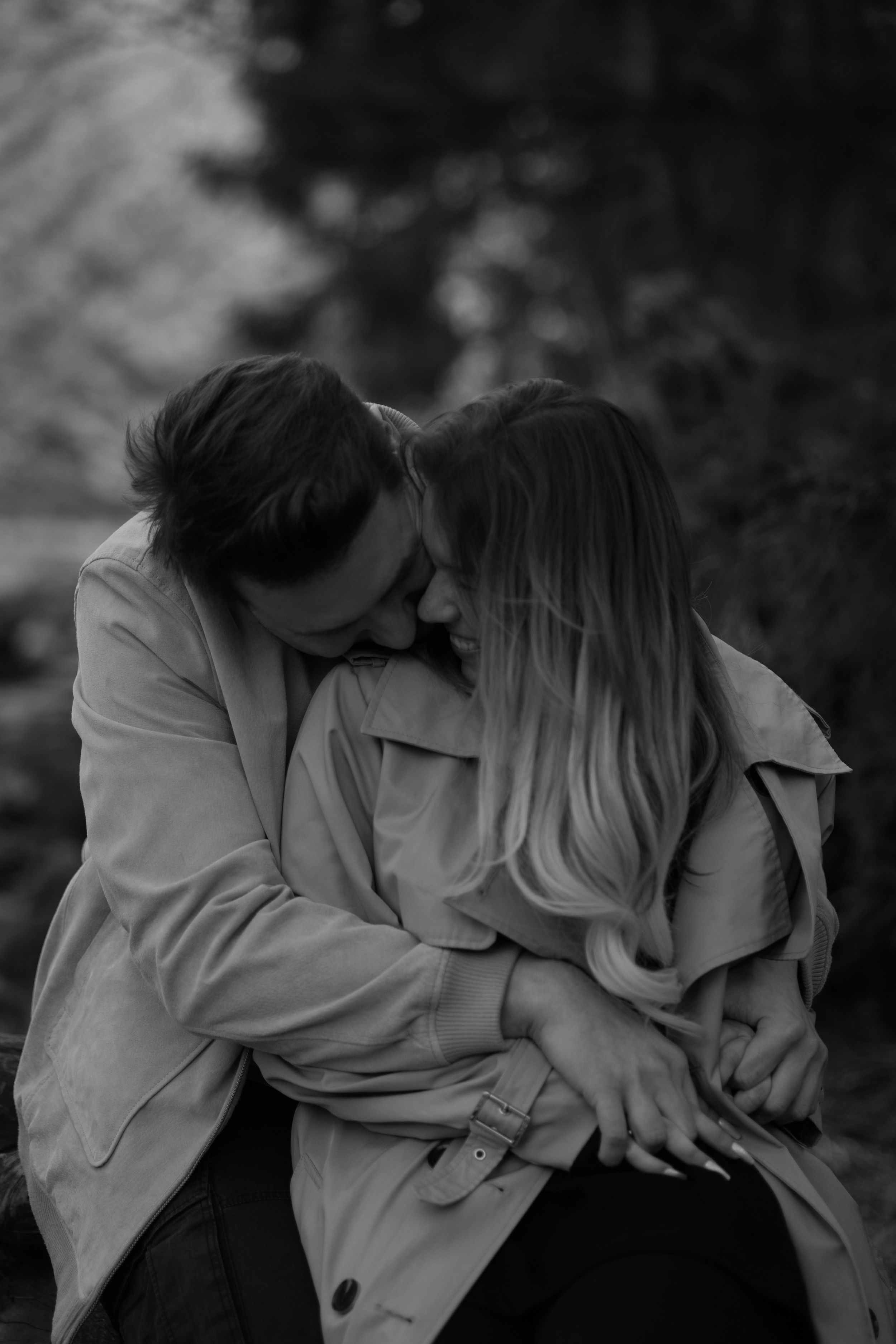 A romantic black and white photo of a couple in trench coats embracing intimately outdoors.