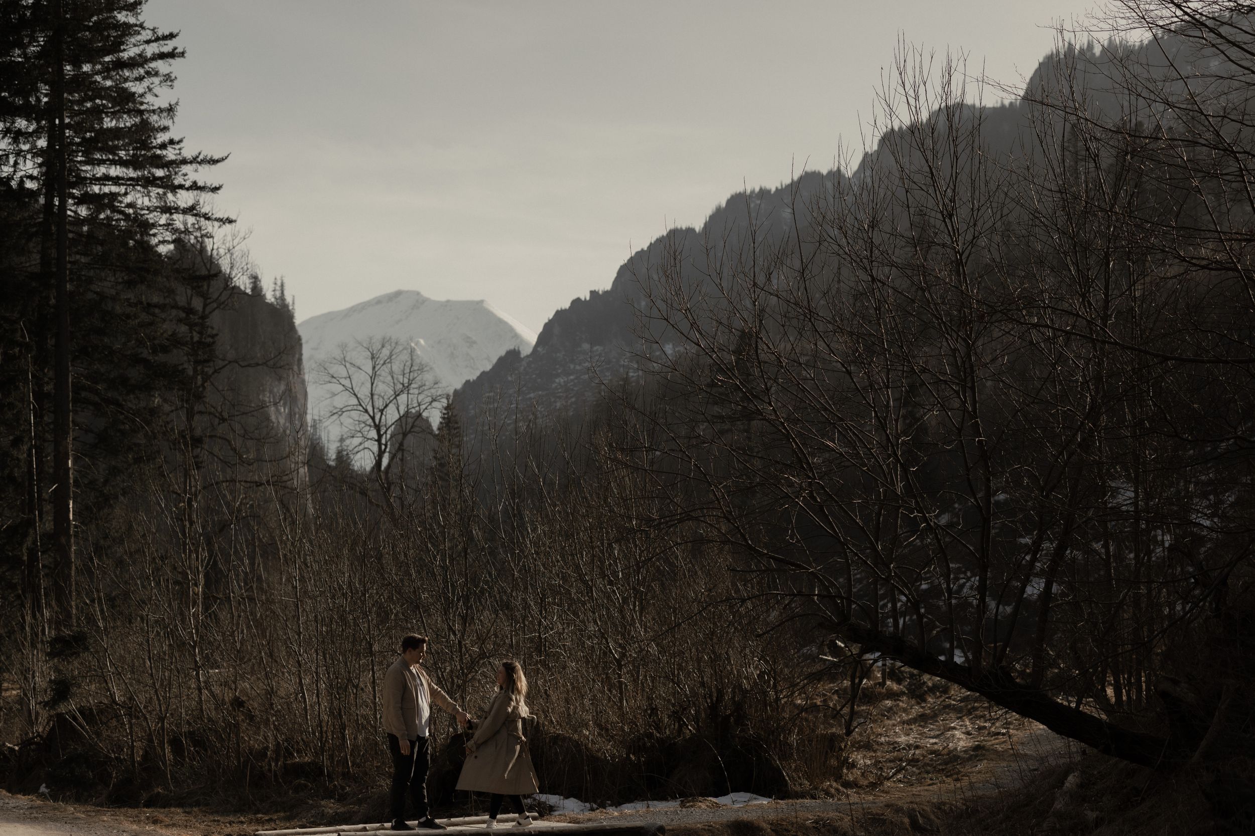 Two people walking together on a mountain path with dramatic peaks and forested slopes in moody overcast lighting.