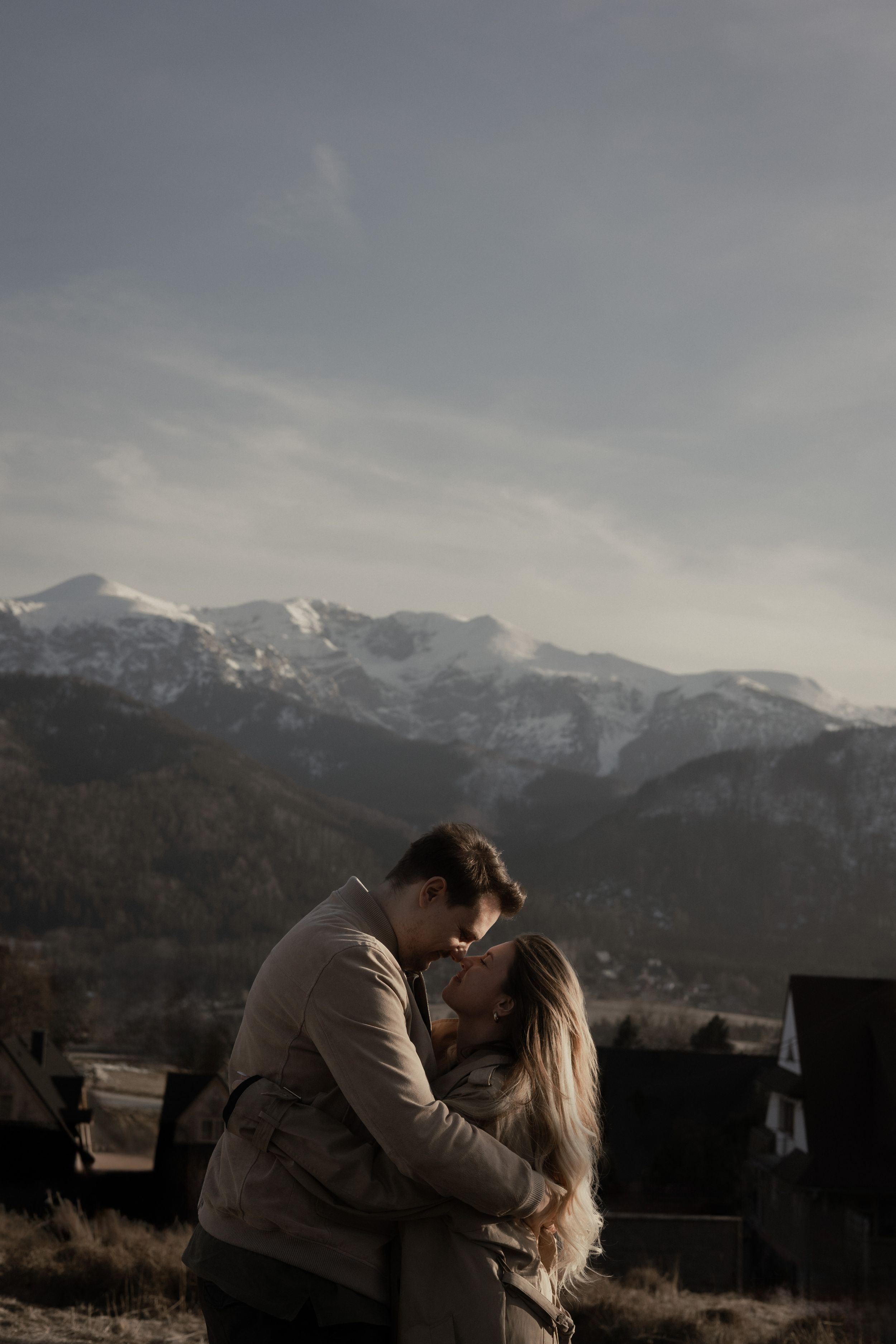 A couple embraces intimately against a backdrop of snow-capped mountains on a moody overcast day.