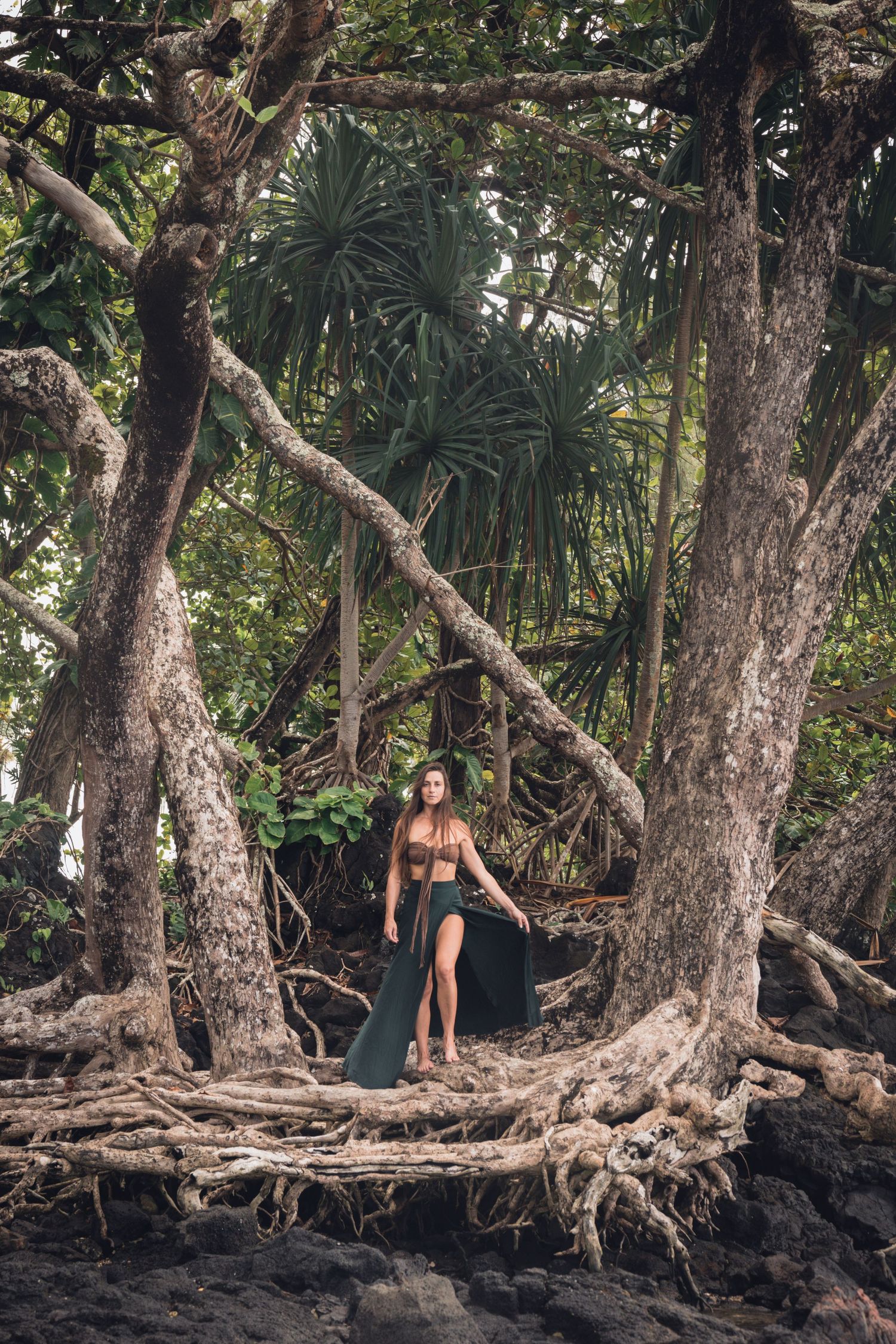 A woman standing on twisted tree roots along a beach with tropical palms and jungle in the background.